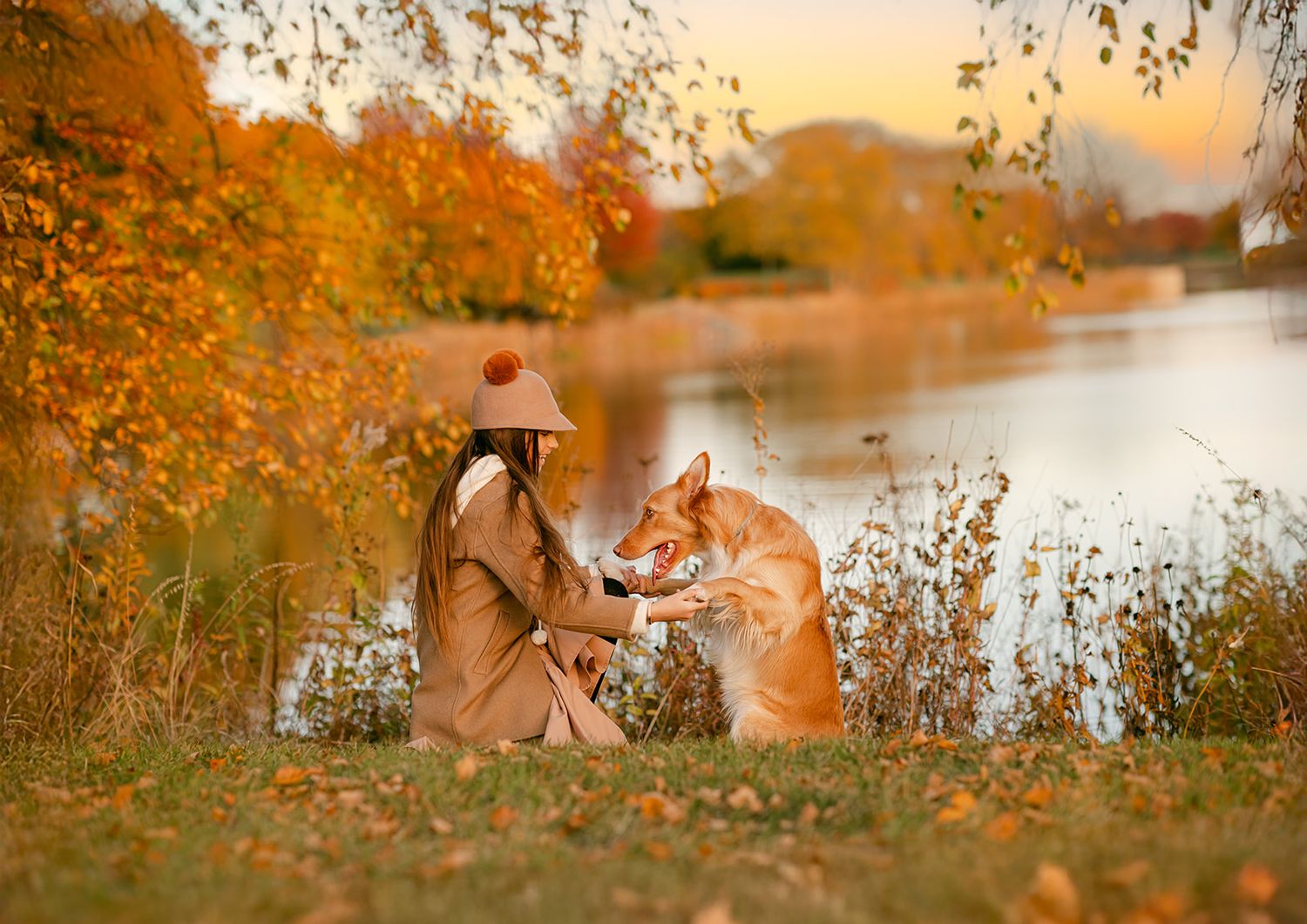 Woman in autumn coat and hat sits by a lake with her golden dog among vibrant fall foliage at sunset.