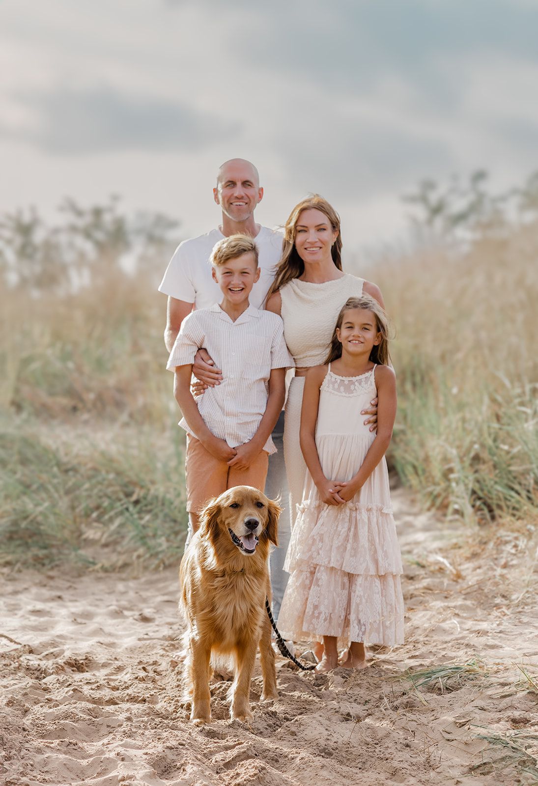 A smiling family of four poses on a sandy beach path with their golden retriever, dressed in coordinating neutral outfits.