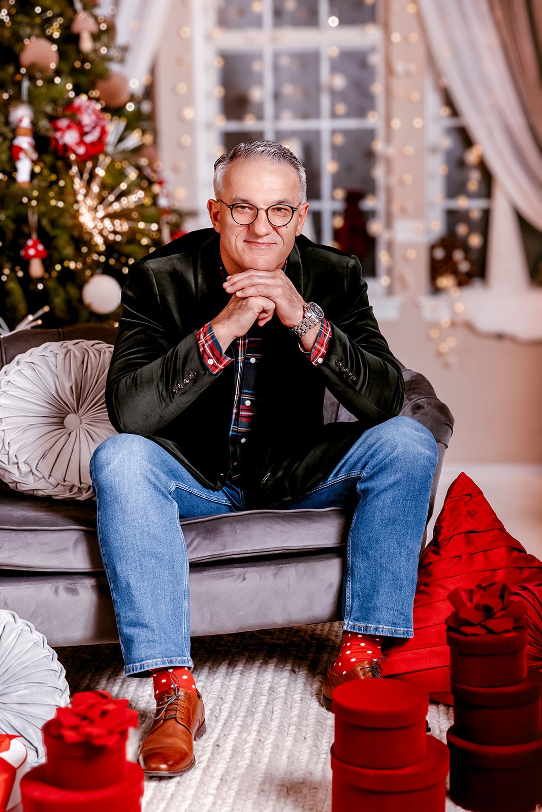 Smiling middle-aged man sitting on sofa holding wine bottle near Christmas tree with red gifts.