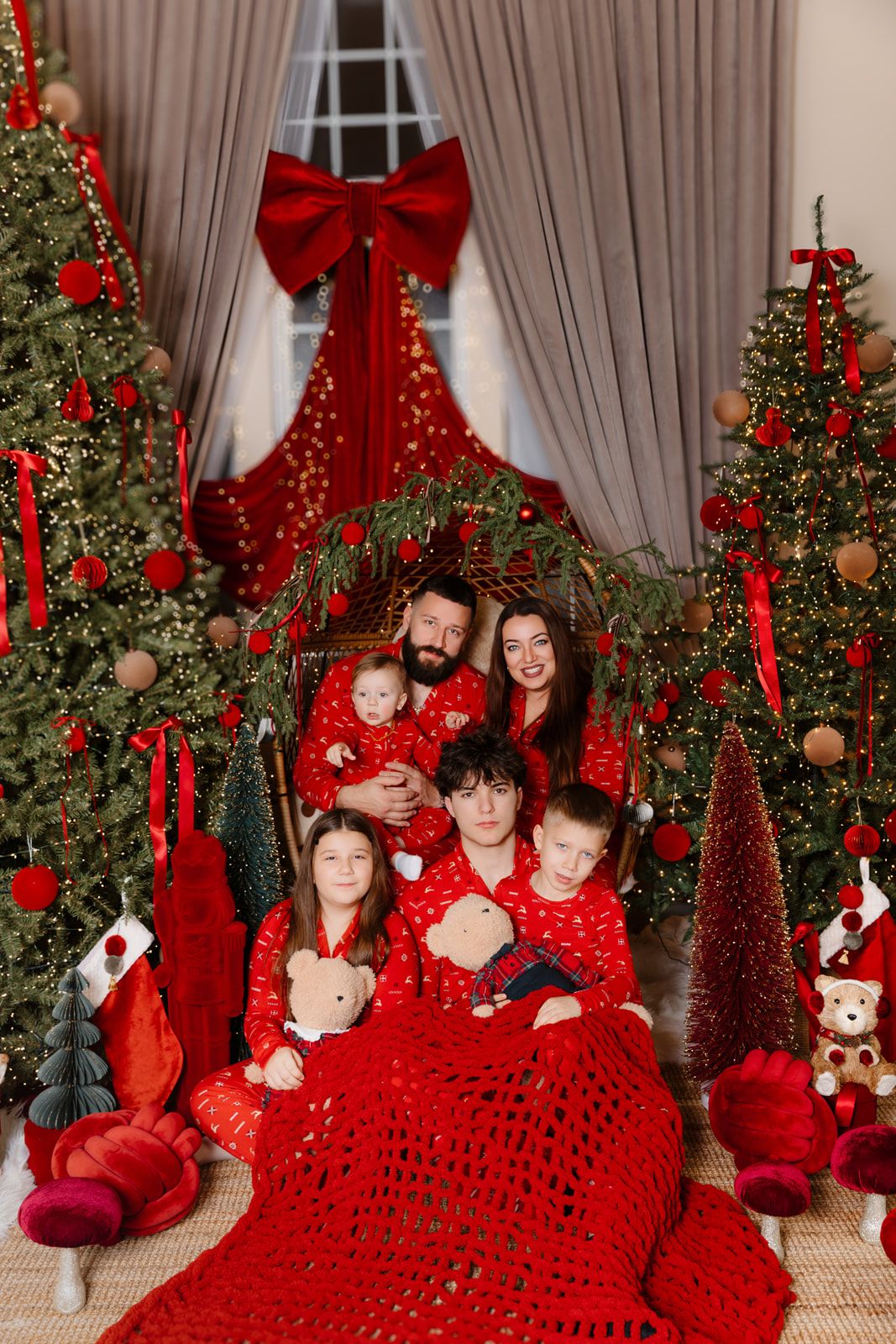 Family in matching red pajamas posing together among decorated Christmas trees with red ornaments and bows.