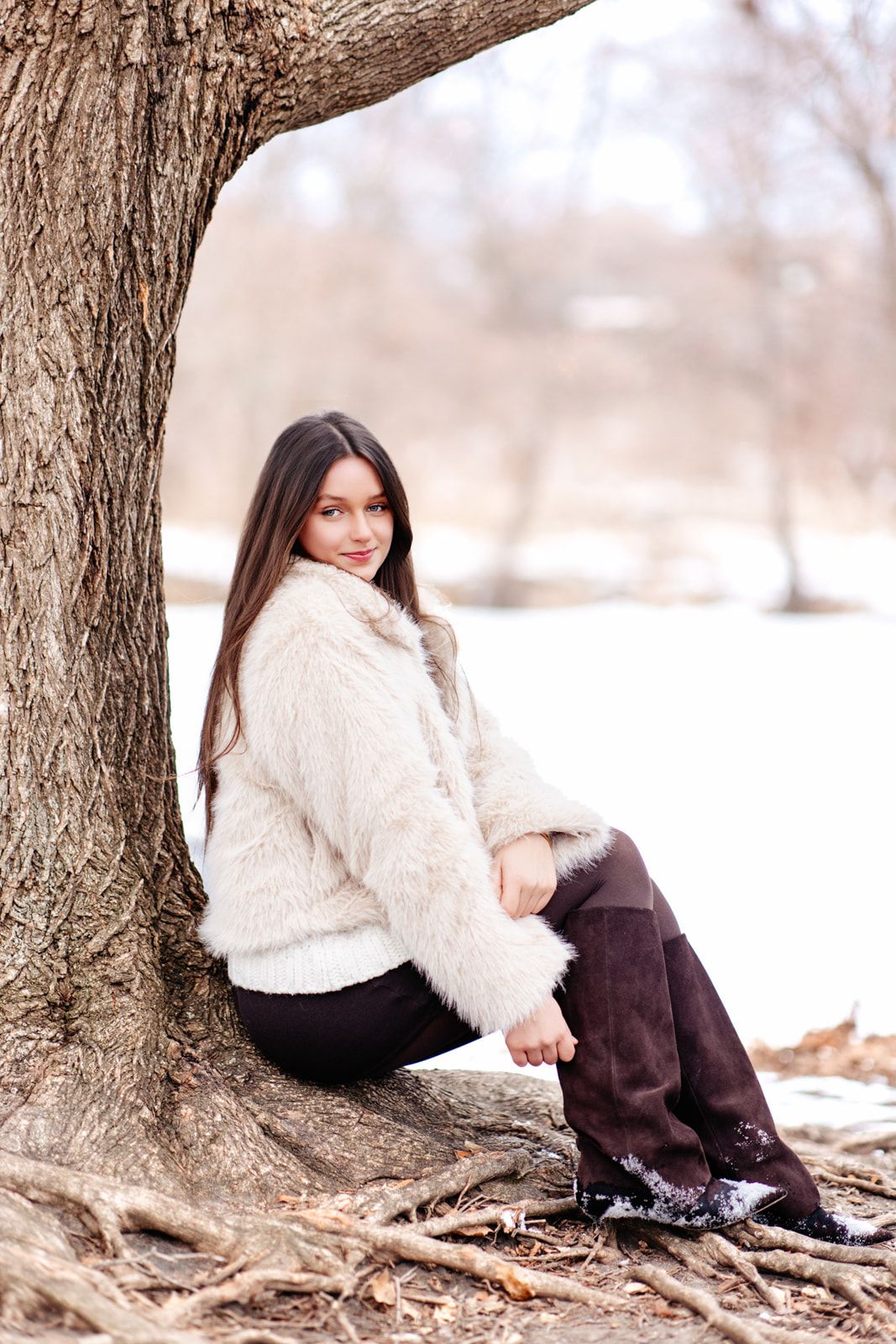 A young woman with long dark hair wearing a cream faux fur coat sits against a large tree in a snowy winter park.