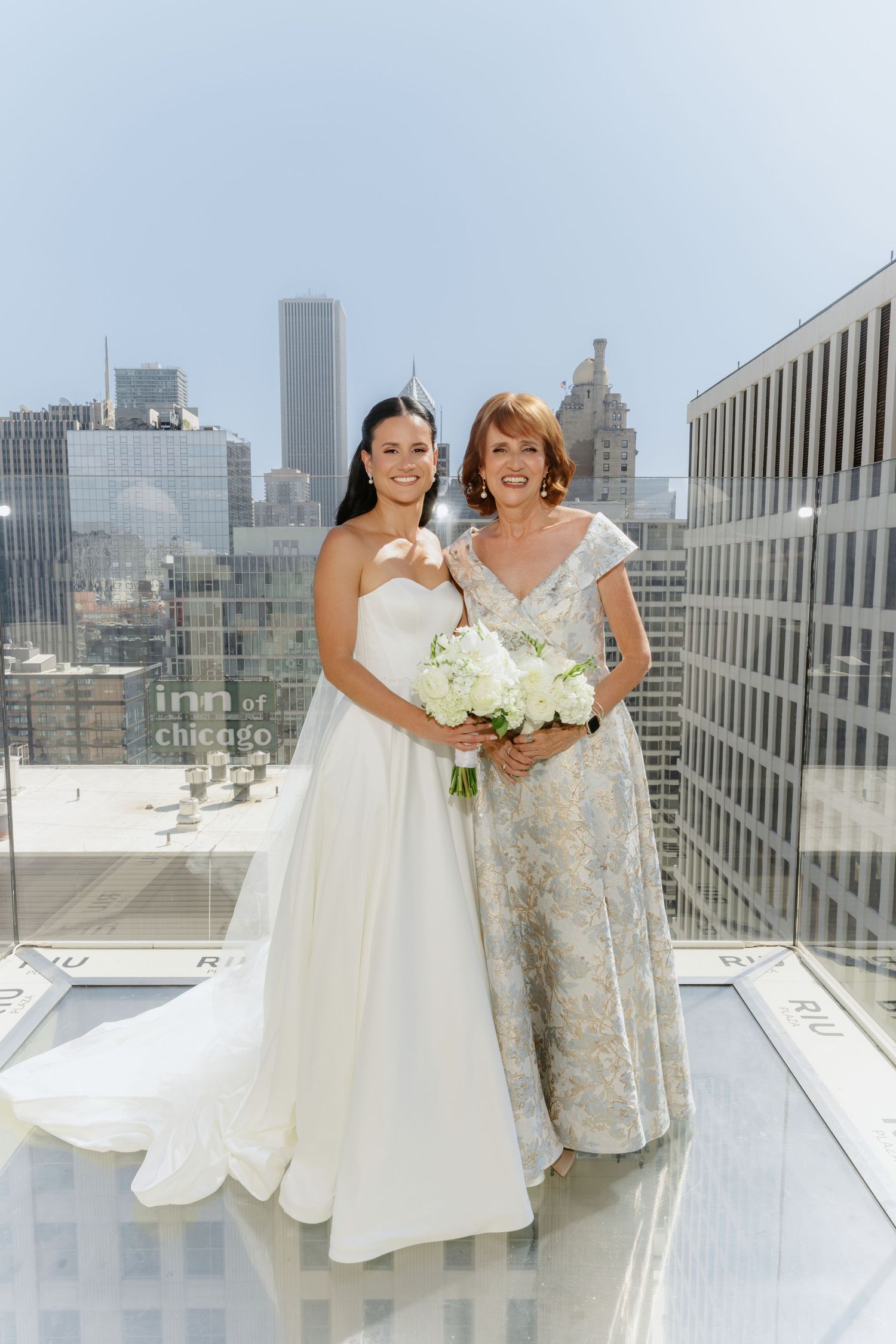 Bride in white strapless gown posing with older woman in floral dress on Chicago rooftop.