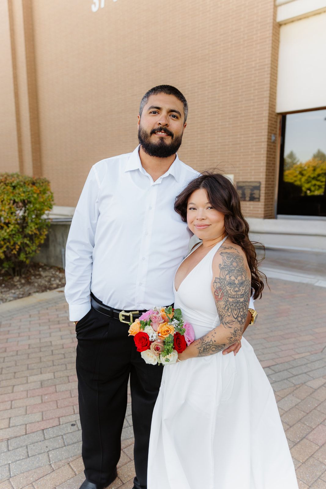 Smiling newlywed couple stands together outside brick building, bride with tattoos holds vibrant floral bouquet.