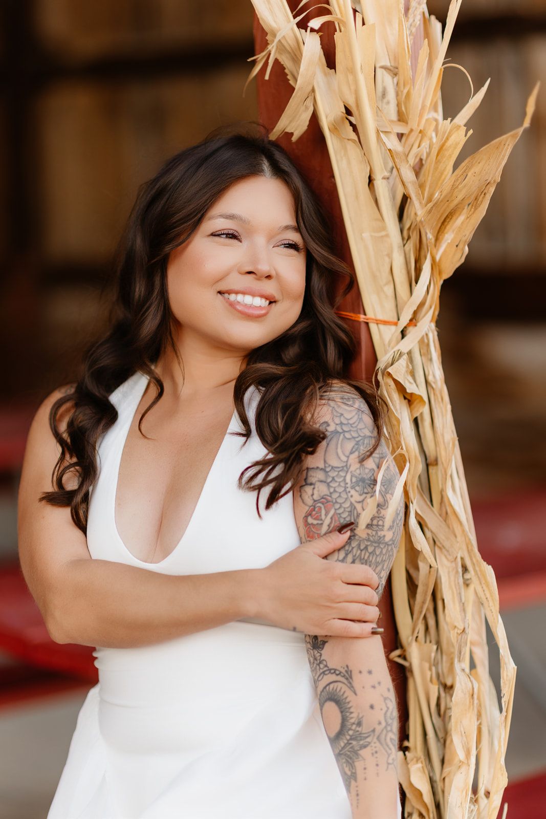 Smiling bride in white halter gown with sleeve tattoo leans against dried corn stalks in barn setting.