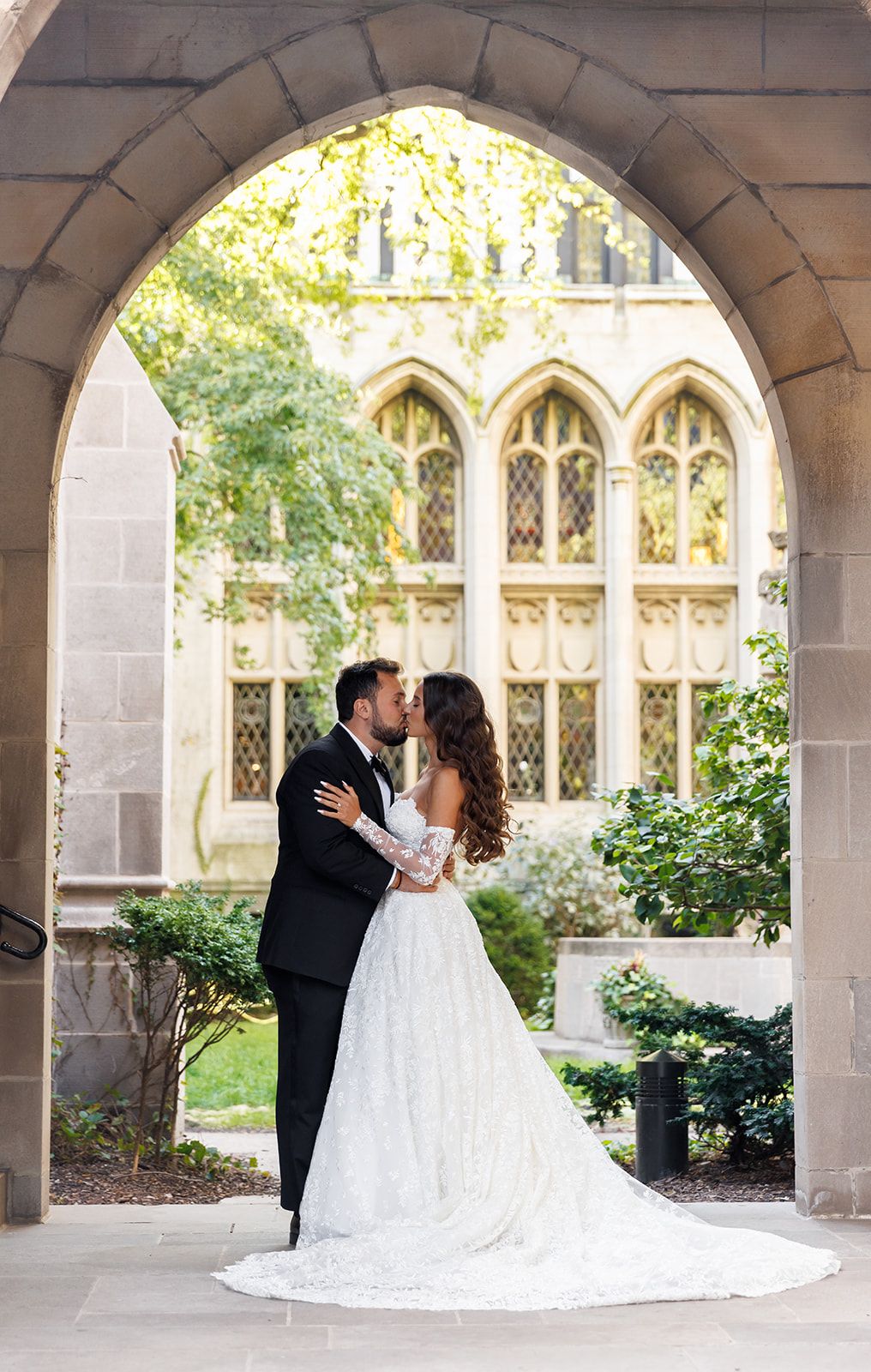 Newlyweds sharing a romantic kiss under an elegant stone archway with Gothic windows in the background.