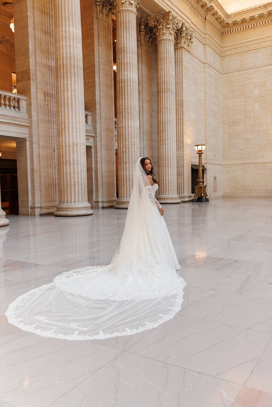 Bride in a white wedding gown with a long lace-trimmed cathedral veil poses in a grand marble hall with tall columns.