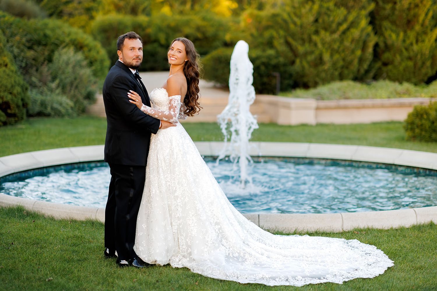 Bride and groom posing elegantly in front of a fountain in a lush garden, bride wearing a stunning lace wedding gown.