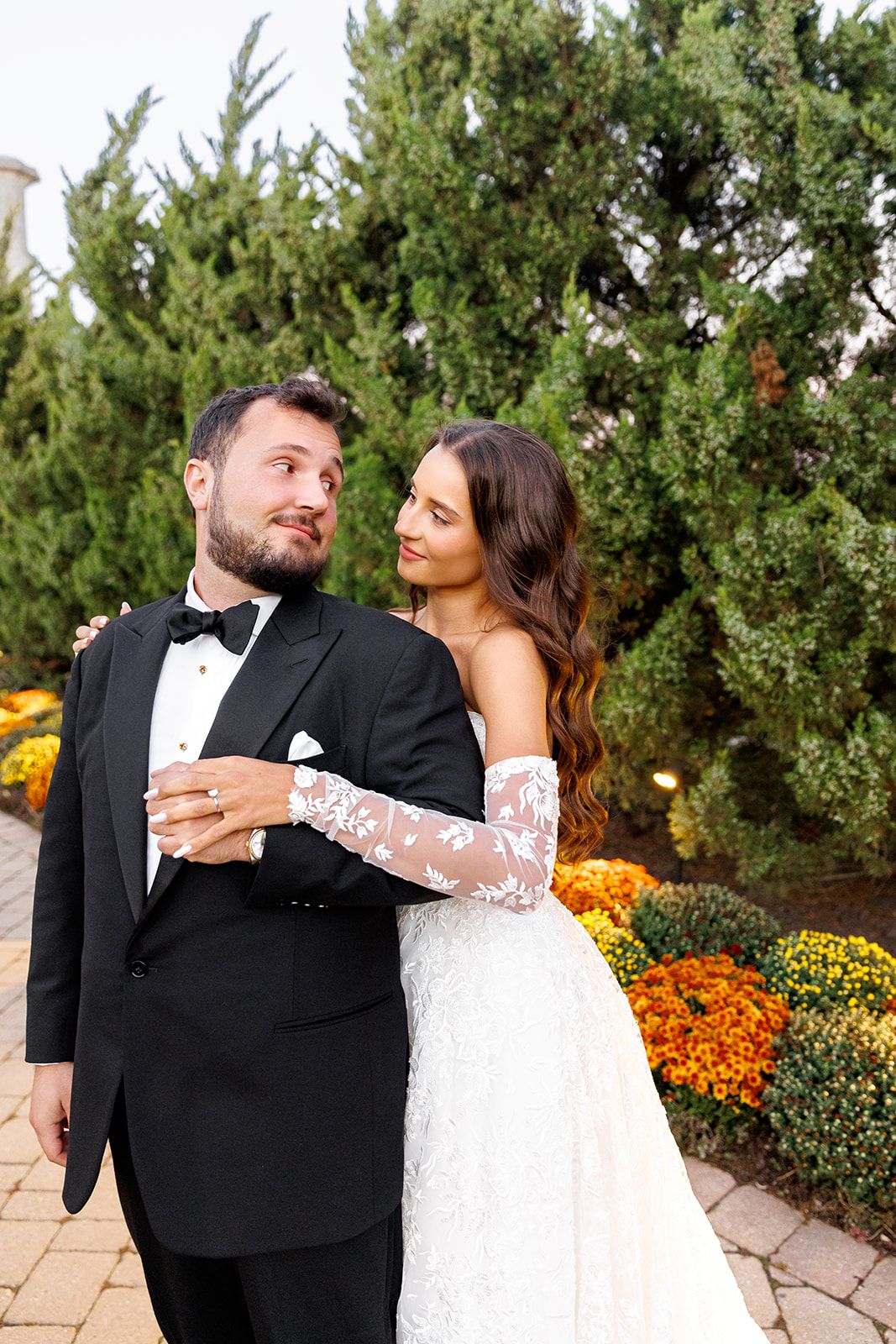 Bride in lace gown embraces groom in black tuxedo outdoors with evergreen trees and fall flowers.