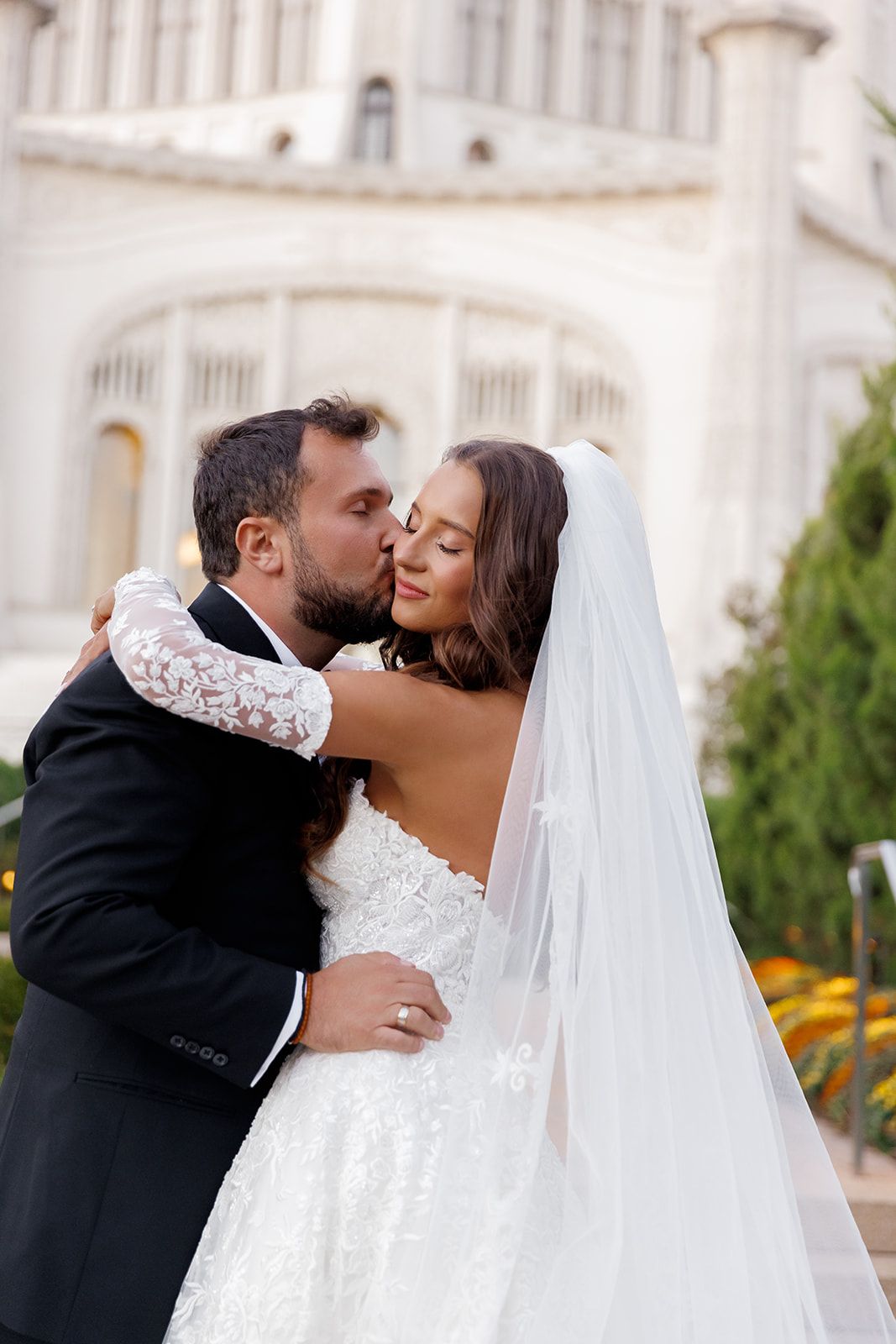 Couple shares a romantic kiss with groom in black tuxedo and bride in strapless lace gown with long veil outdoors.