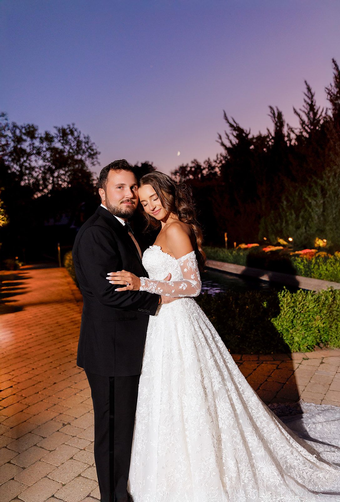 Smiling bride and groom pose closely together on a brick pathway under a beautiful purple sunset sky.