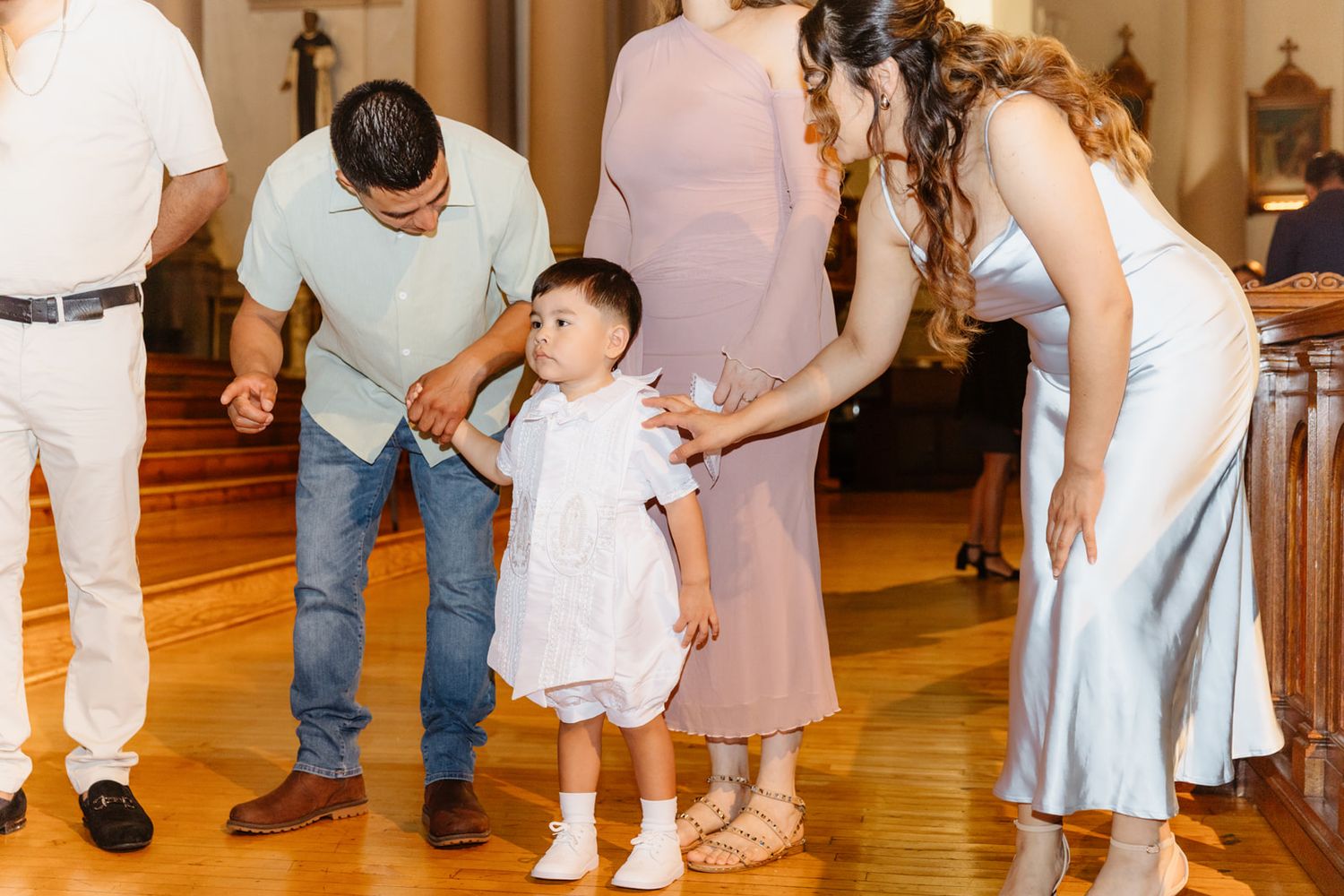Family members help a toddler in white outfit walk on wooden church floor during a baptism celebration.