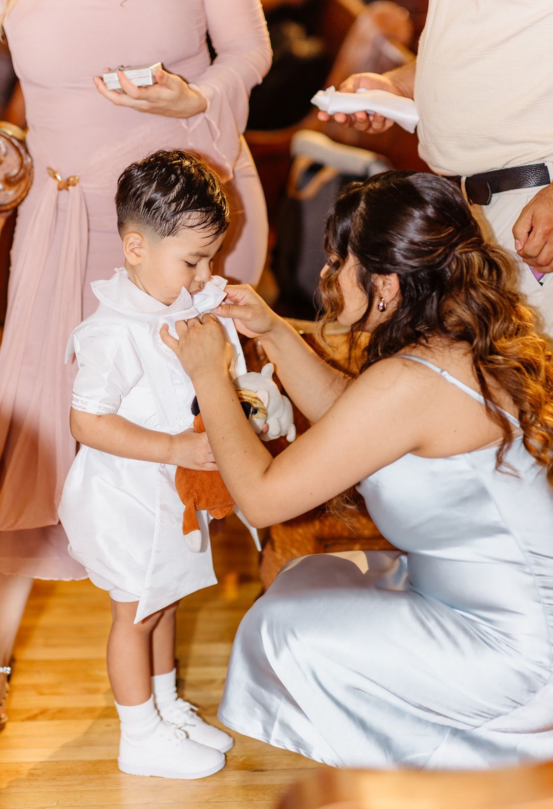 A woman in a light blue dress kneels to wipe a young boy's nose during a baptism celebration.