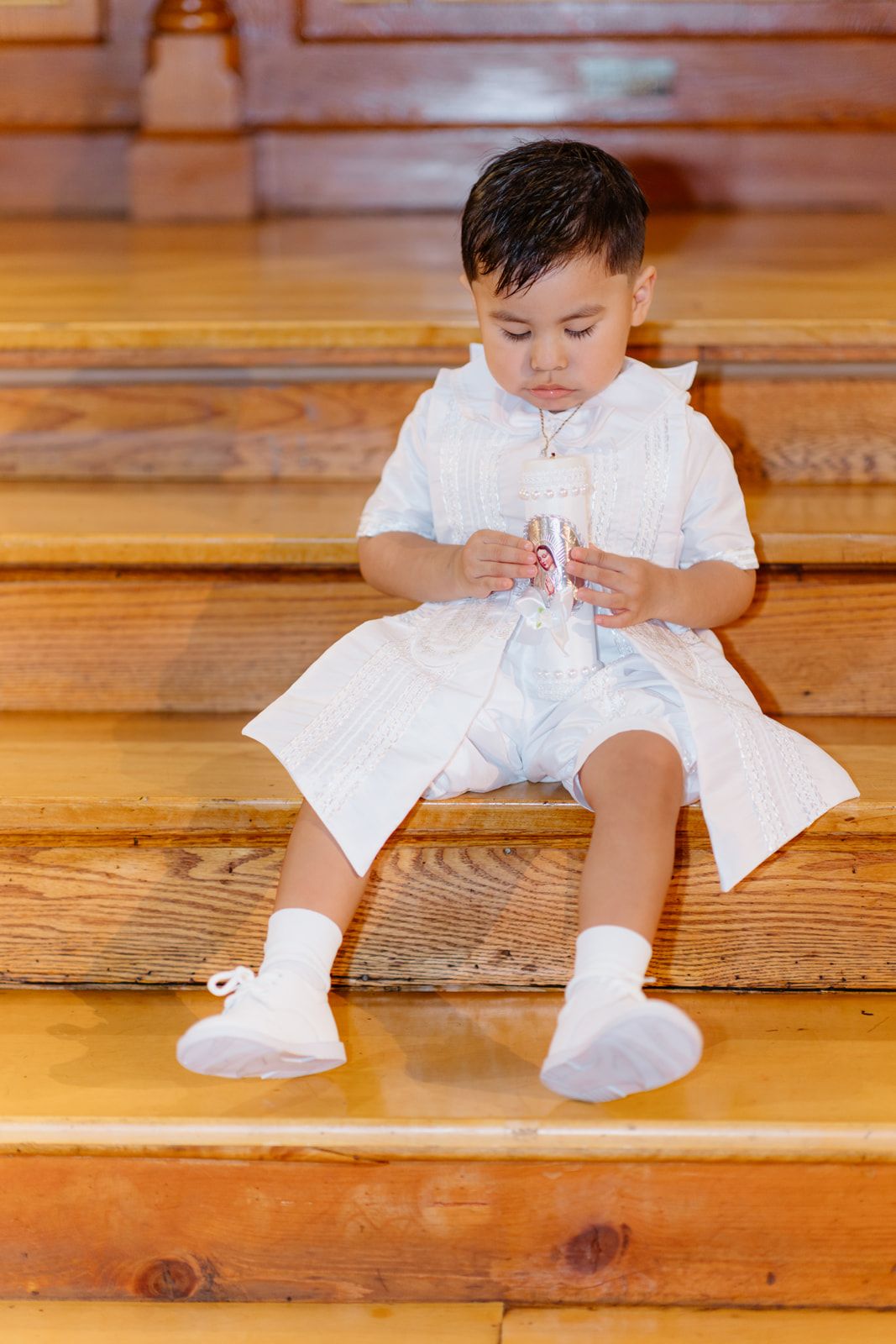 Toddler boy dressed in white christening clothes sits on wooden steps examining a decorative candle.