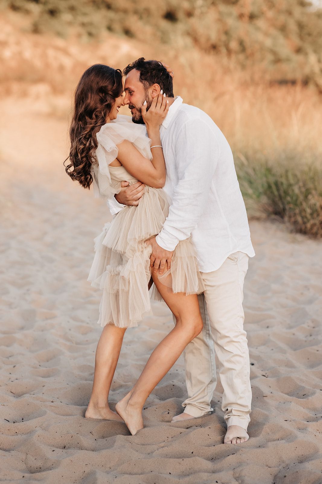 Romantic couple embracing on sandy beach at golden hour, woman in beige tulle dress, man in white shirt.