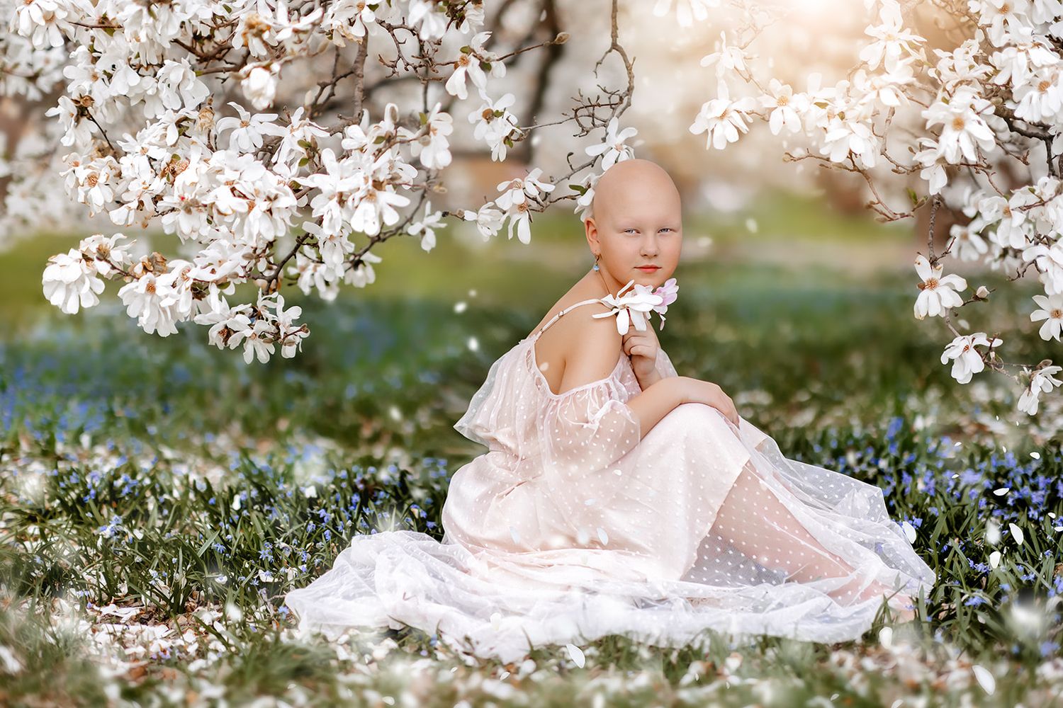 A bald young girl in a white lace dress sits among blooming magnolia trees, holding a white flower in sunlight.