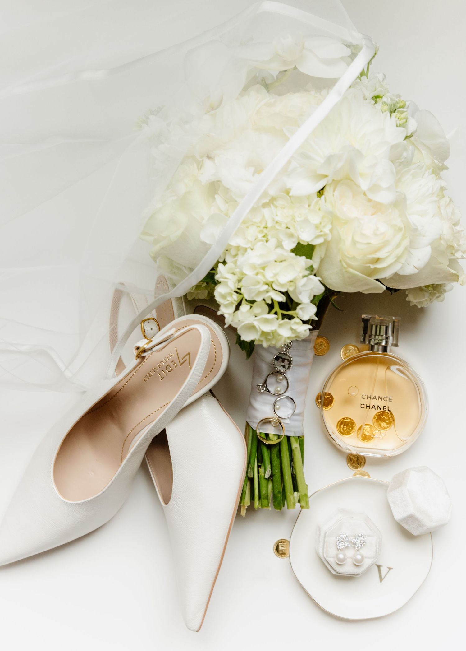 Elegant bridal flat lay featuring white heels, Chanel perfume, wedding rings, and a lush white floral bouquet on a white background.