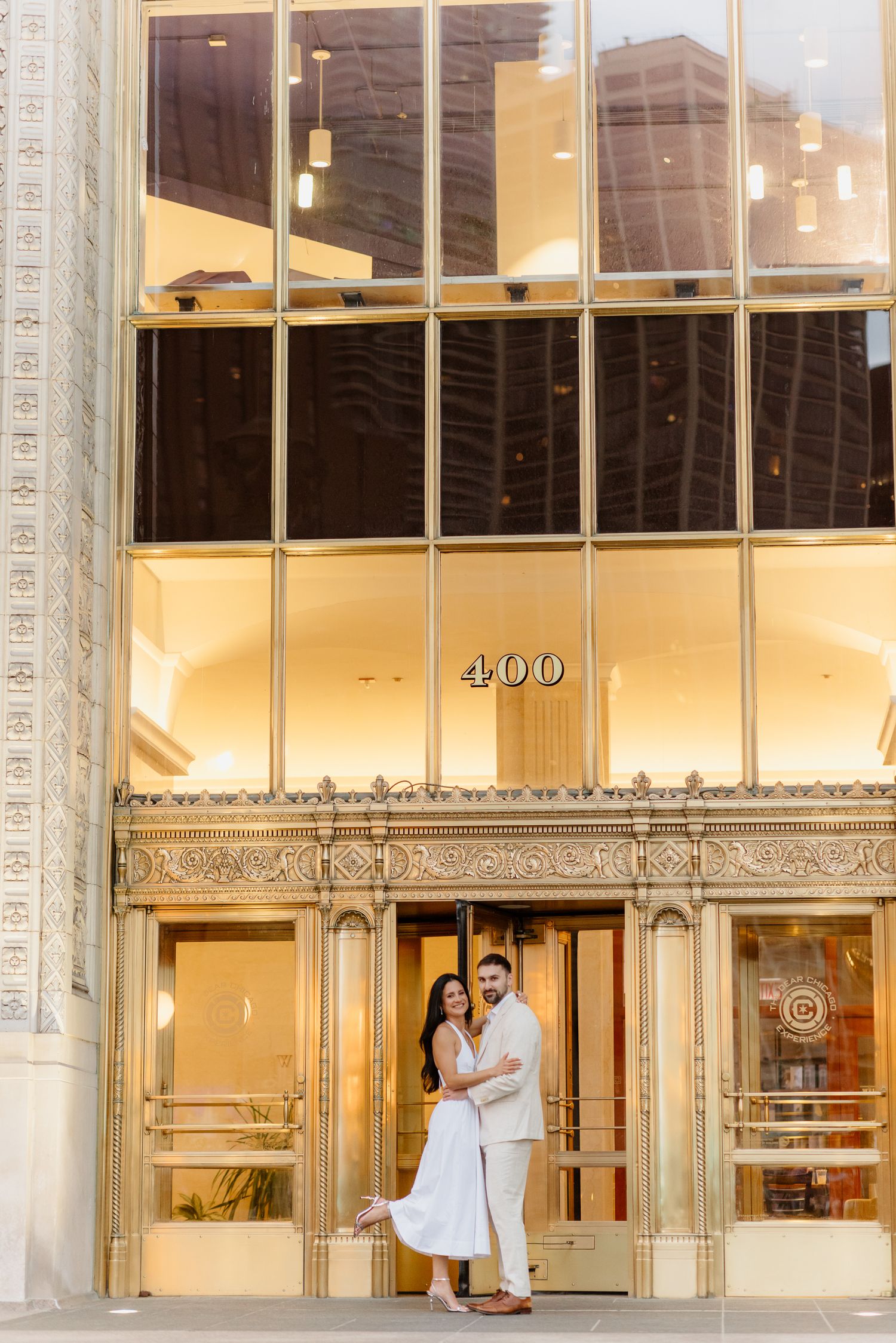 Couple in white outfits embracing outside 400 building entrance with ornate gold details in urban setting.