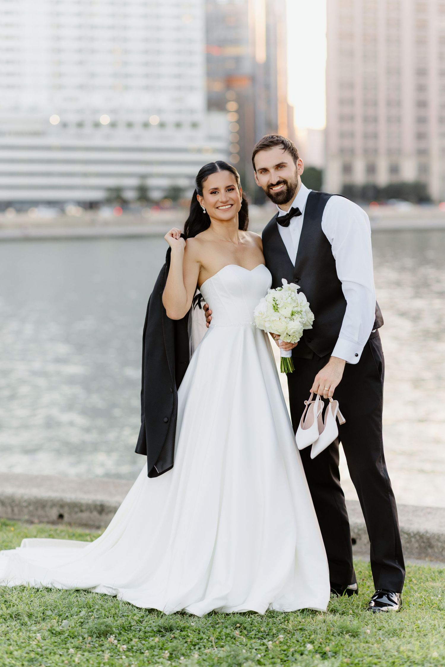 Bride in white strapless gown holds bouquet as groom in black vest and bow tie carries her heels by a city waterfront.