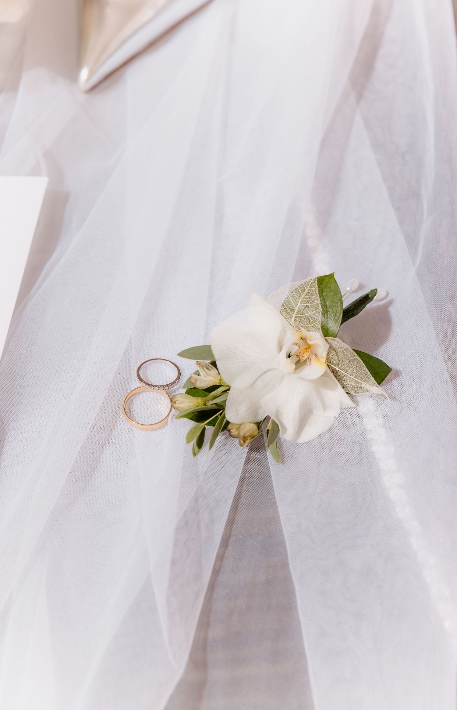 Two gold wedding rings placed on a white orchid and greenery boutonniere draped over a white bridal veil.