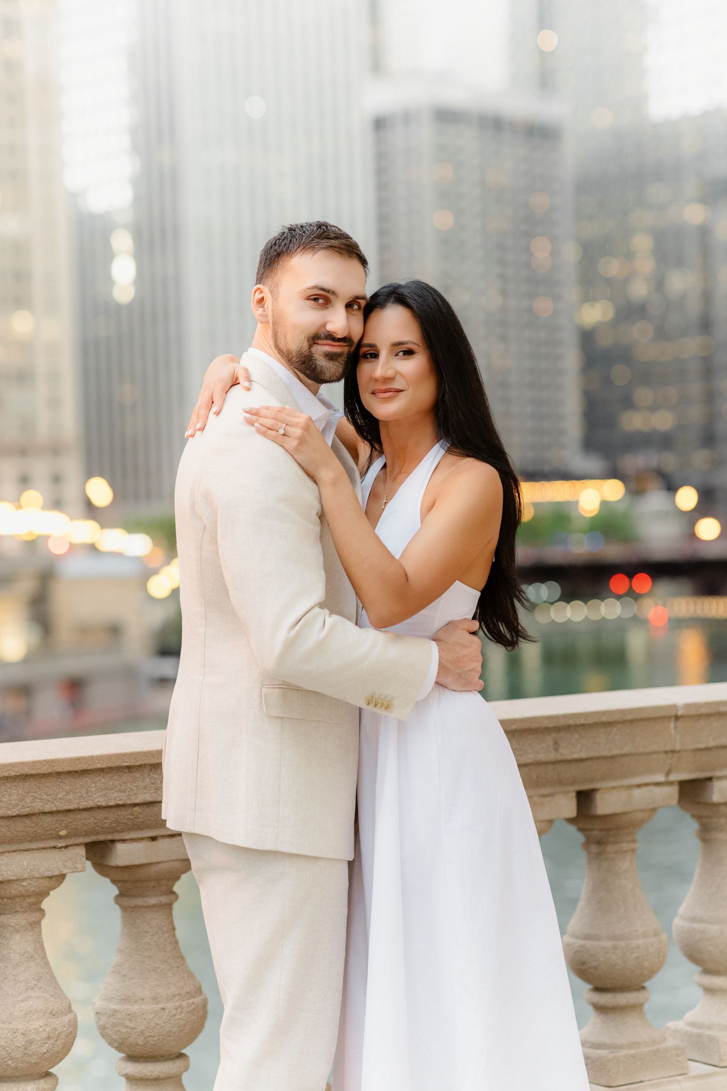 Couple in white outfits embracing on a balcony with Chicago River and skyline behind them.