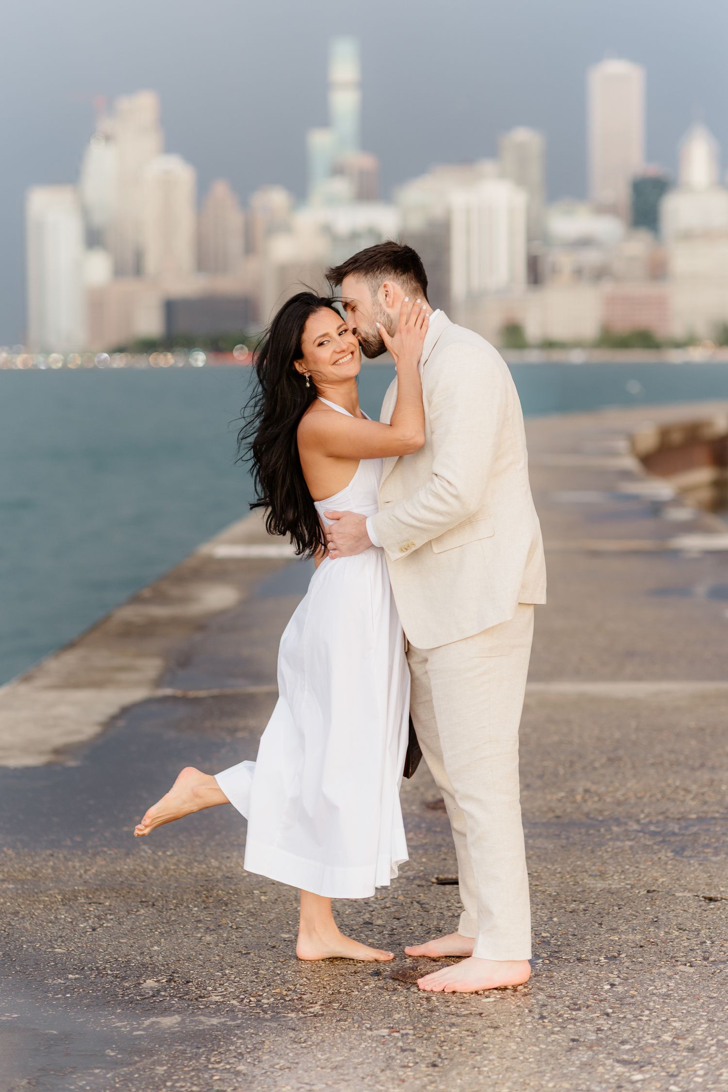 Smiling woman in white dress lifts leg as couple embraces barefoot on Chicago lakefront pier.