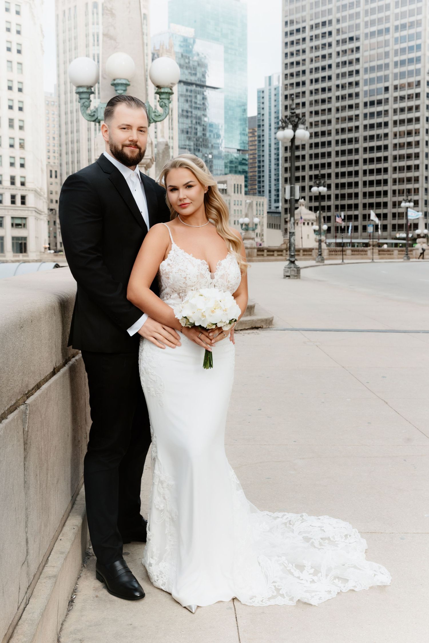 Bride in fitted lace gown and groom in black tuxedo pose together with Chicago skyline in background.