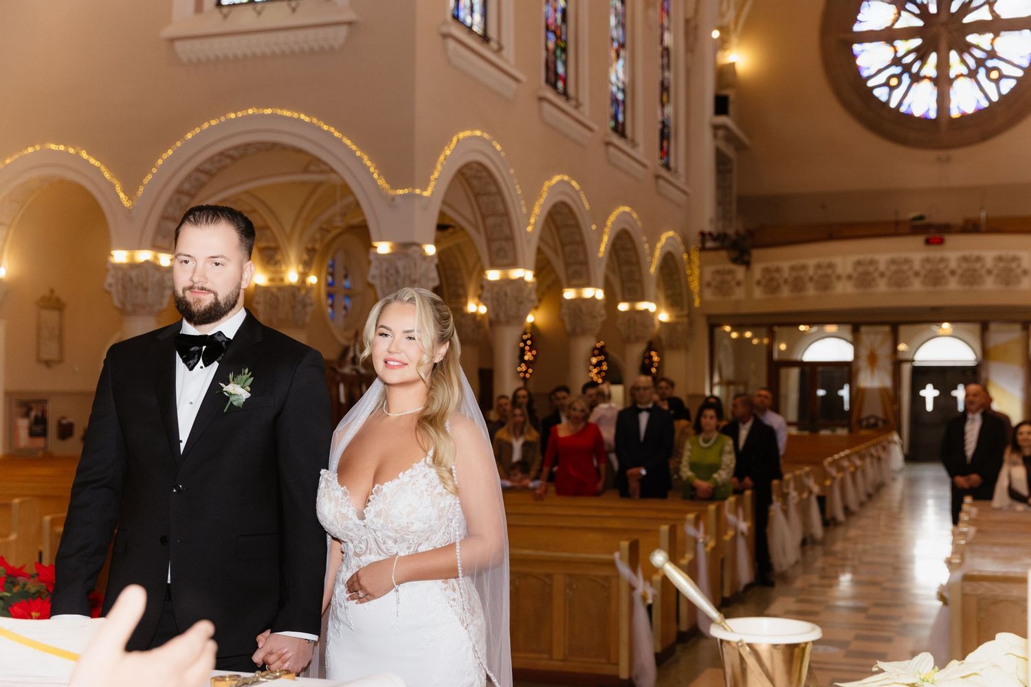 Smiling bride and groom hold hands while standing at church altar with guests and rose window visible behind.