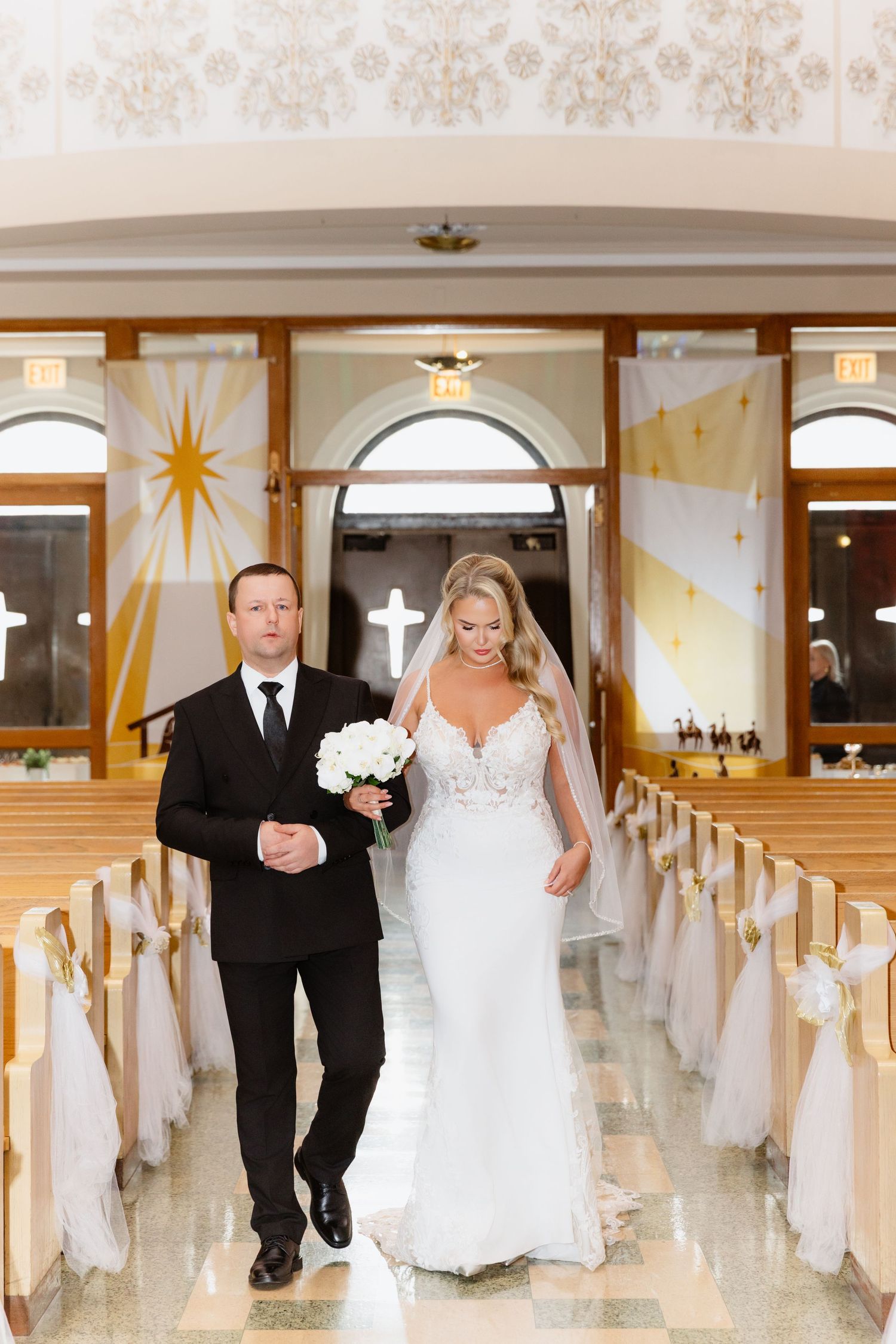 A bride in a white lace gown and veil walks down the aisle escorted by a man in a black suit.