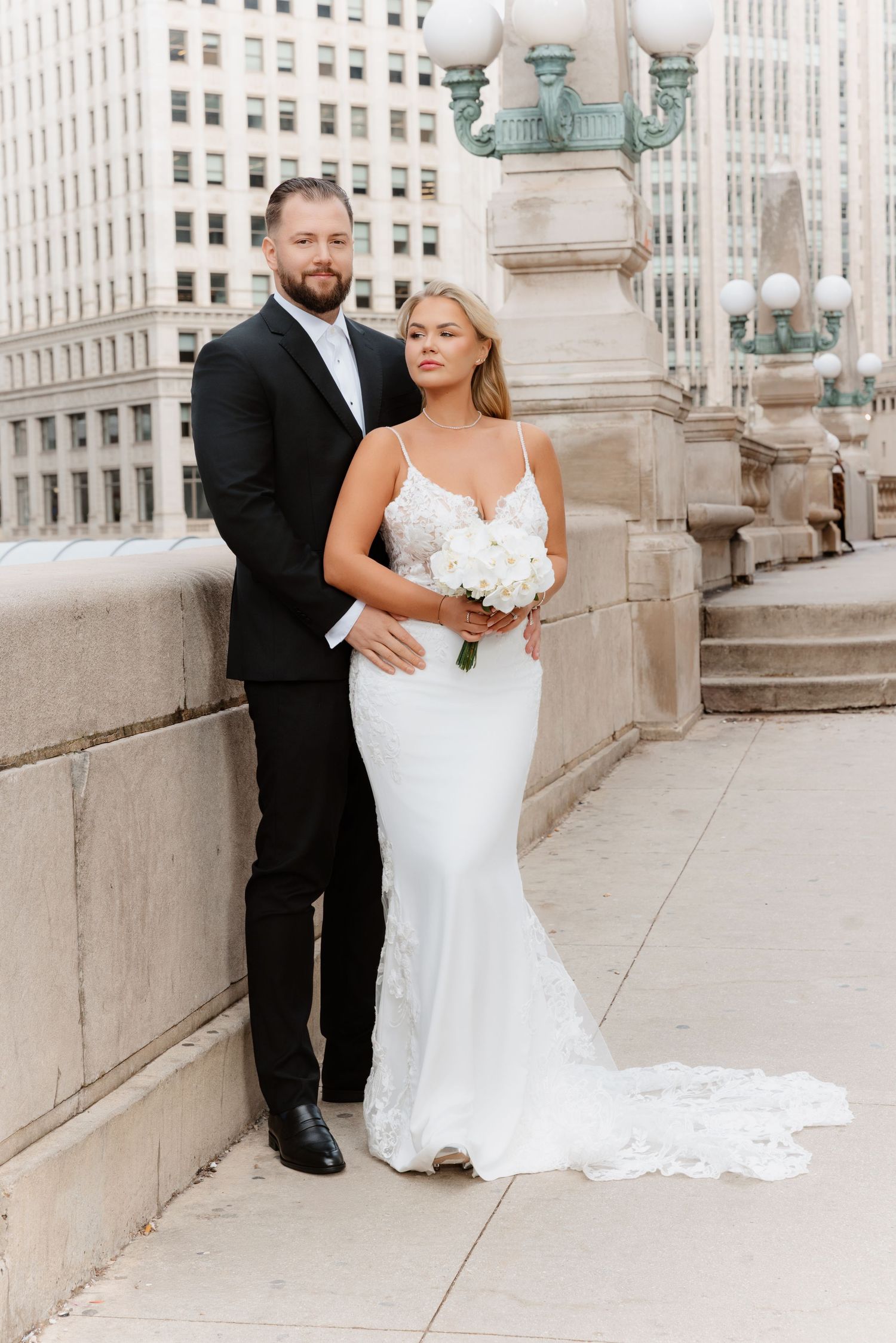 Bride in fitted lace gown with train poses with groom beside stone column on Chicago bridge.