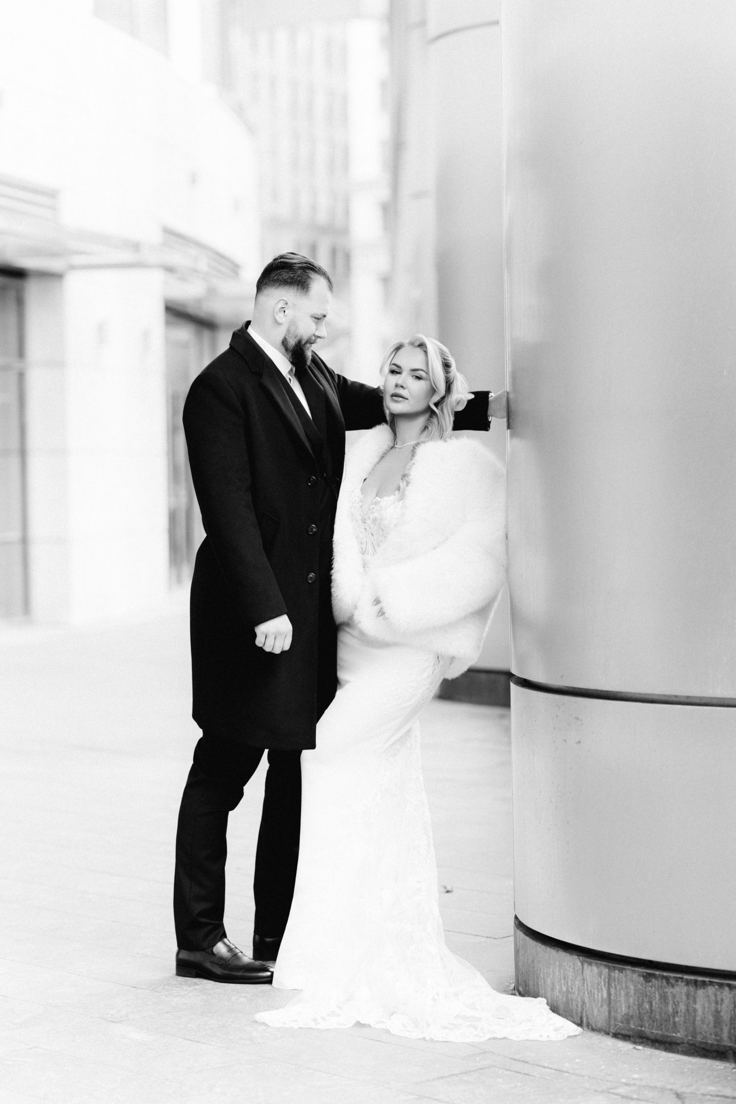 Black and white photo of bride in fur wrap and groom in black coat by urban columns.