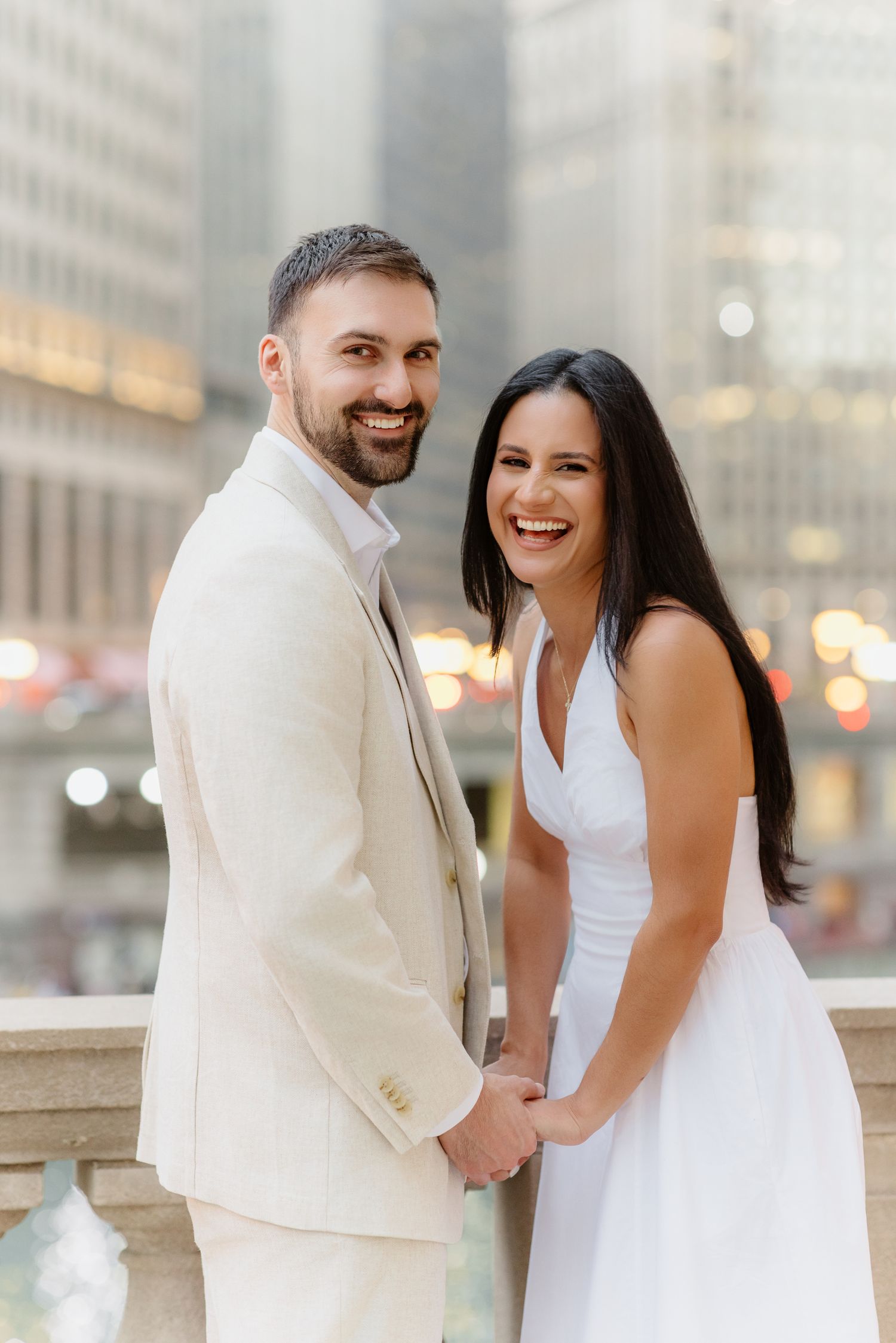 Joyful couple in white and cream attire holds hands laughing with city bokeh lights behind them.