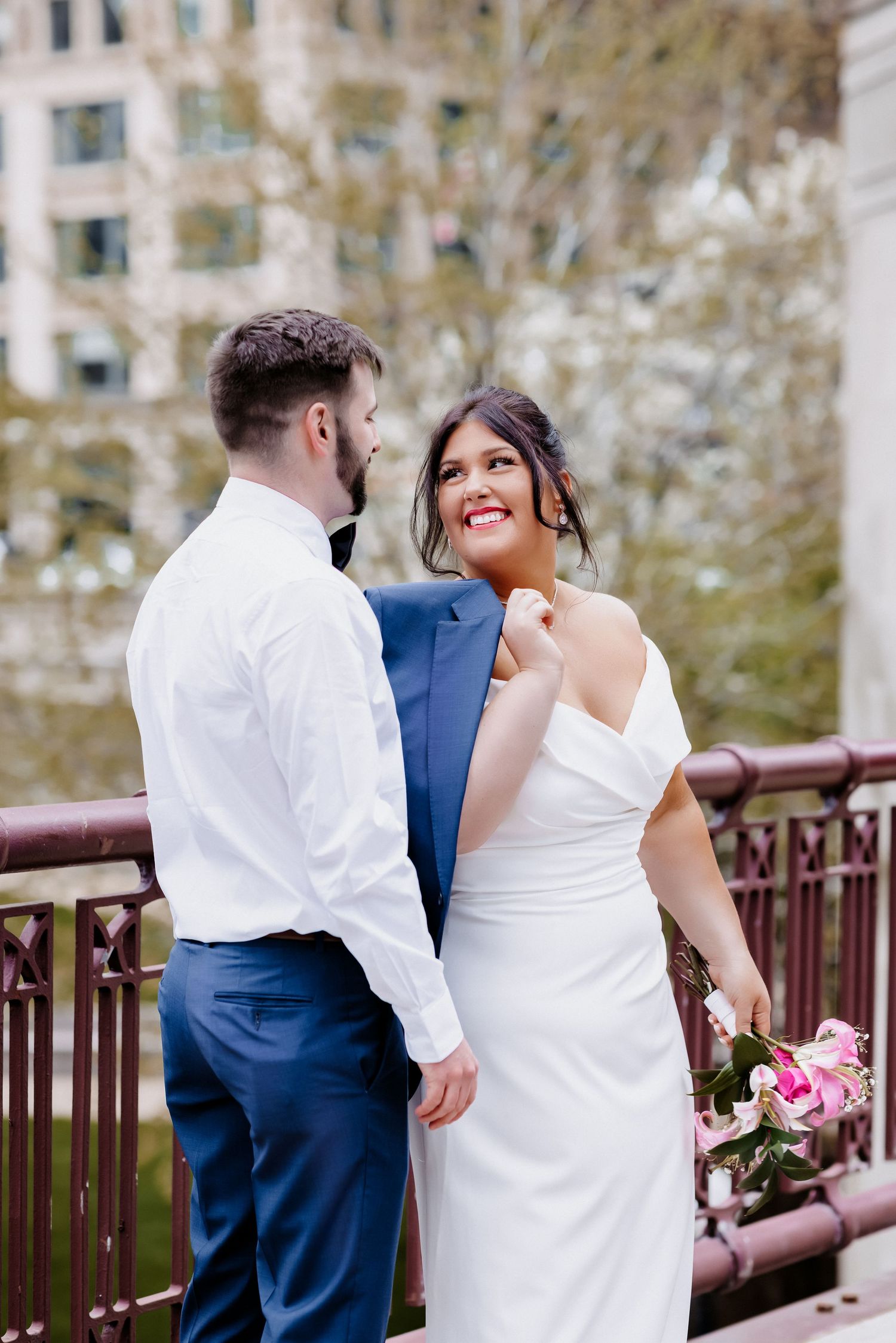 Bride in white gown holds grooms blue jacket and smiles at him on a decorative bridge with spring blossoms.