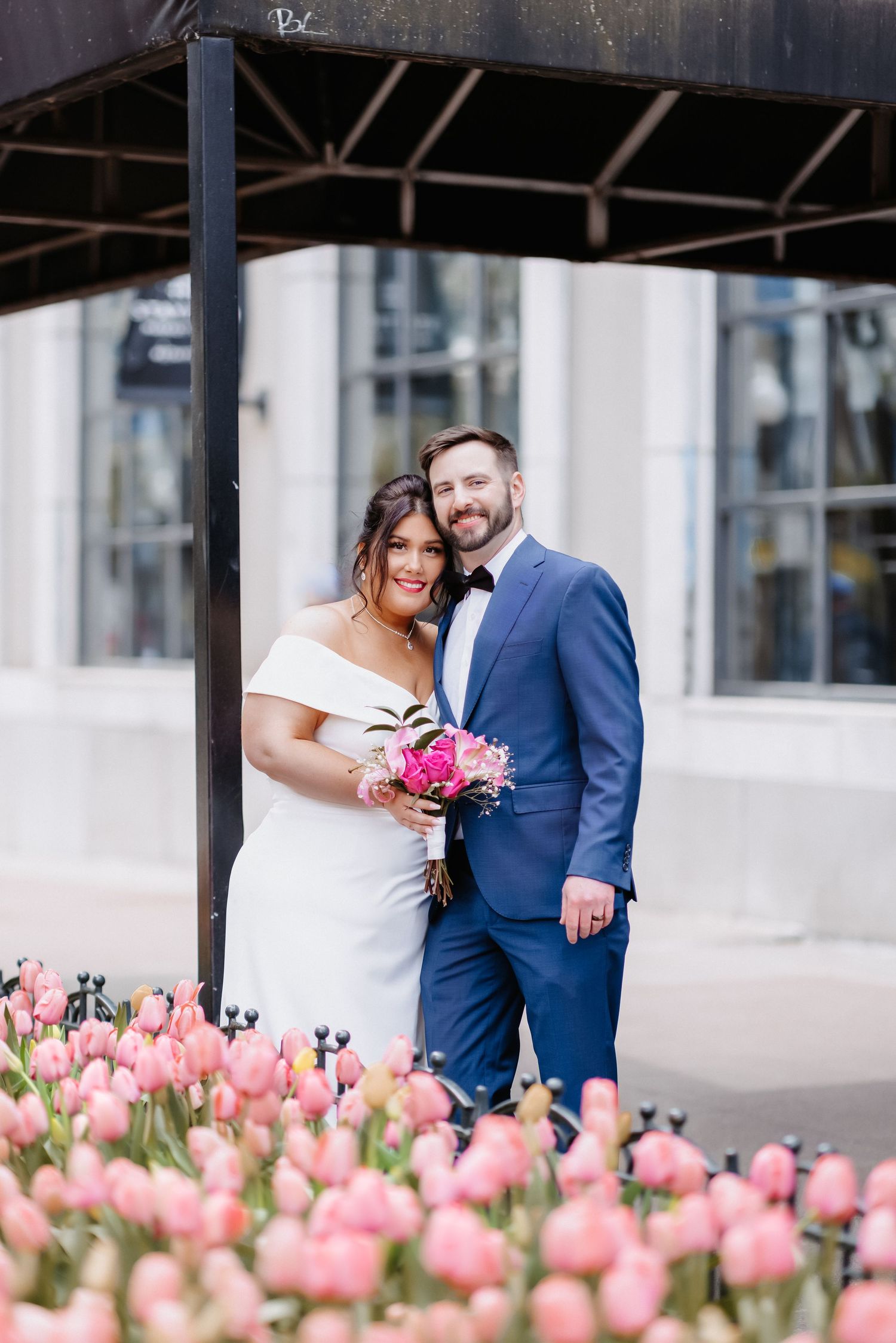 Wedding couple smiles together surrounded by pink tulips, bride in white gown holding pink bouquet, groom in navy suit.