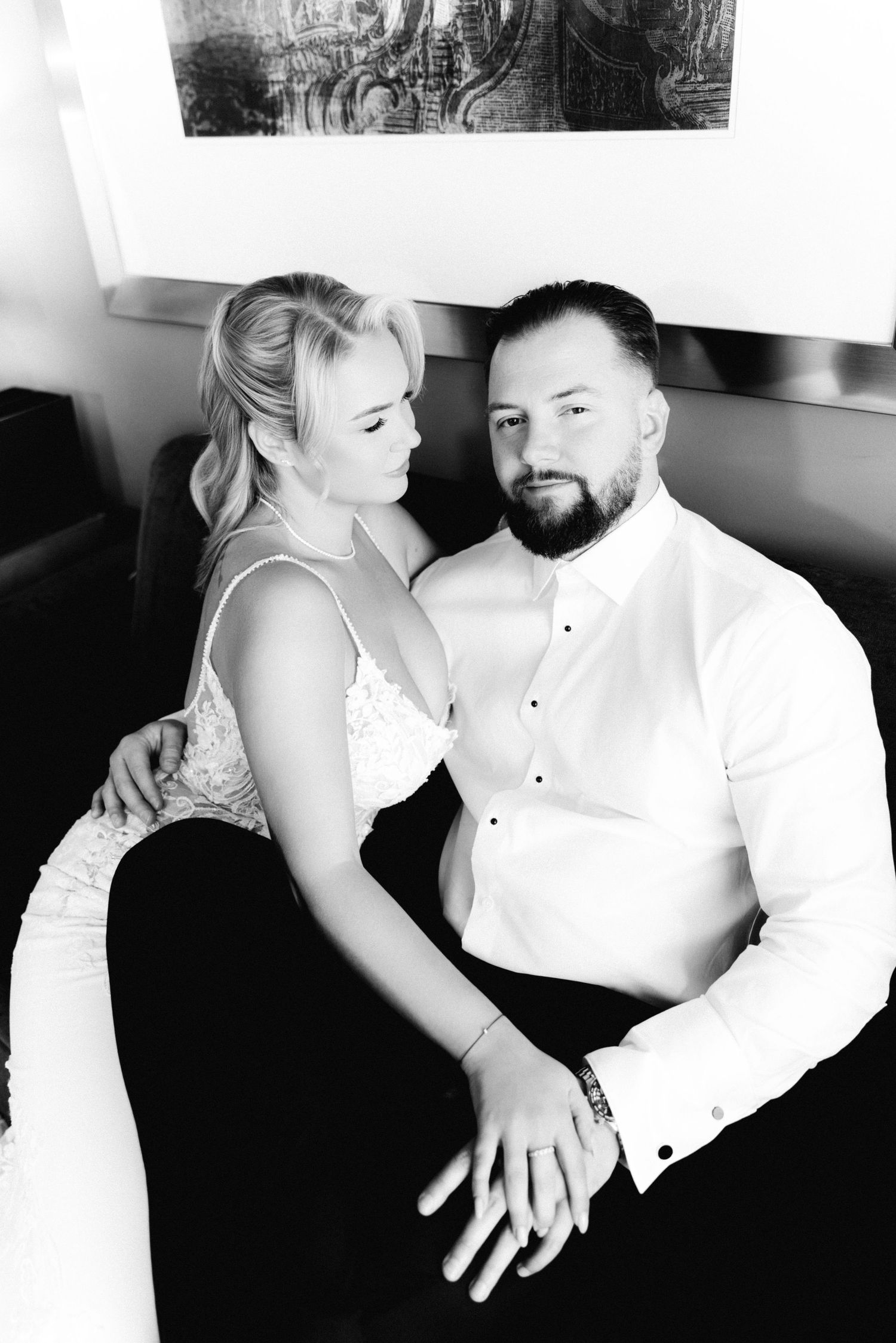 Black and white portrait of bride looking at bearded groom while sitting together on dark sofa.