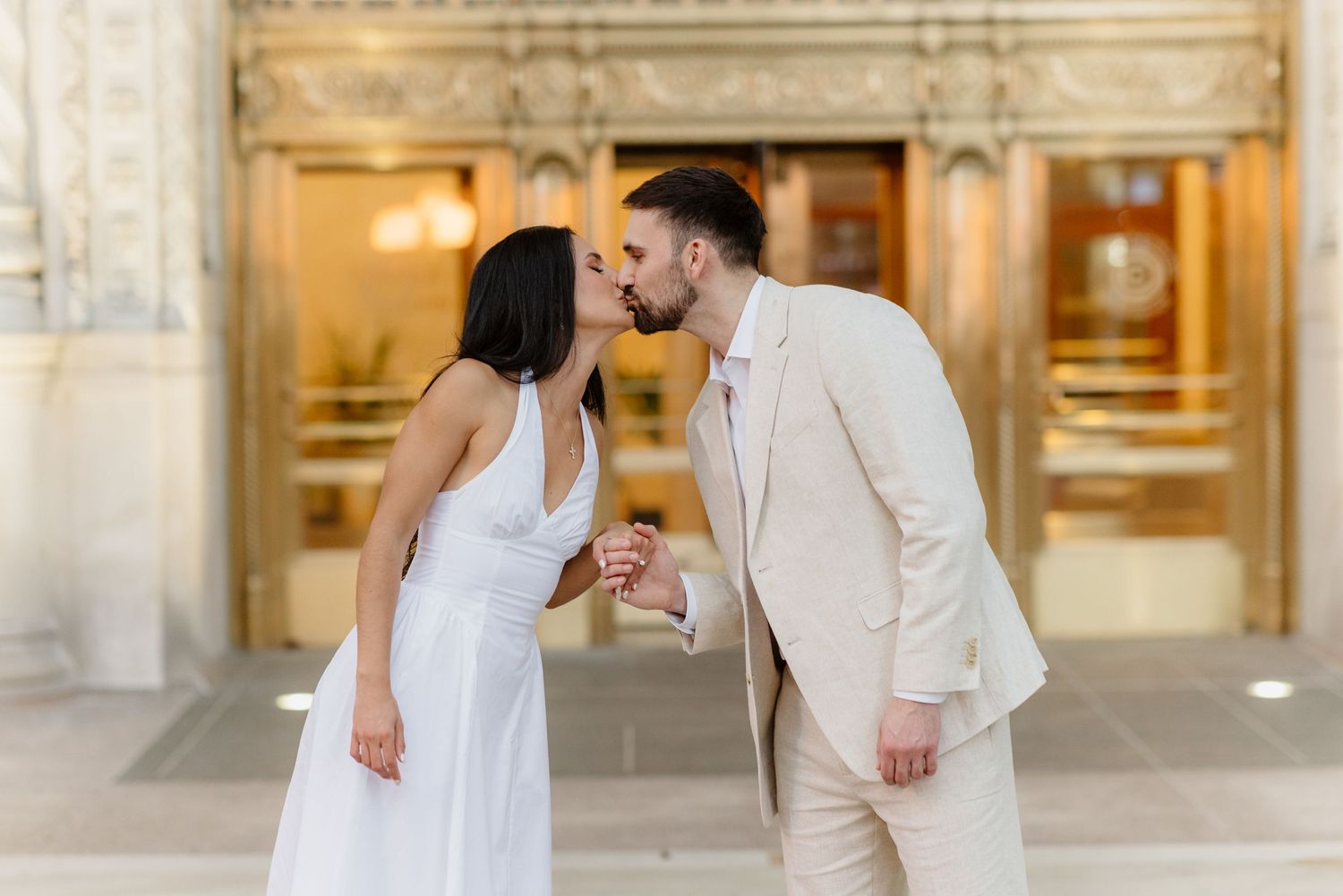 Couple sharing romantic kiss outside ornate gold building entrance in coordinating outfits.