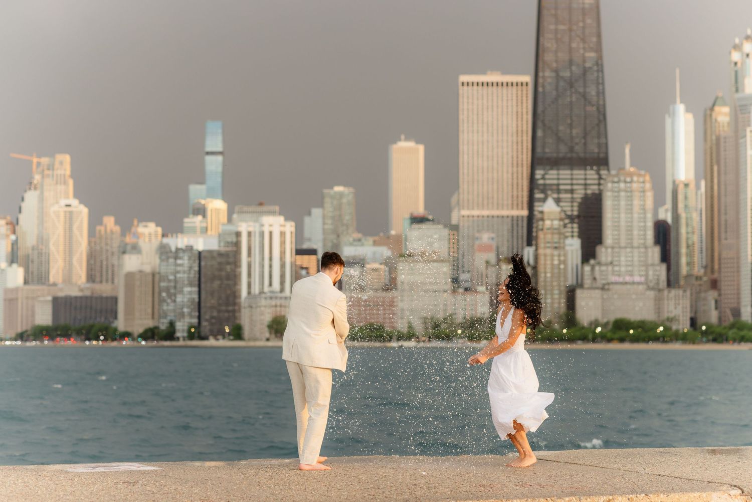 Woman in white dress running toward partner on Chicago lakefront with skyline at sunset.