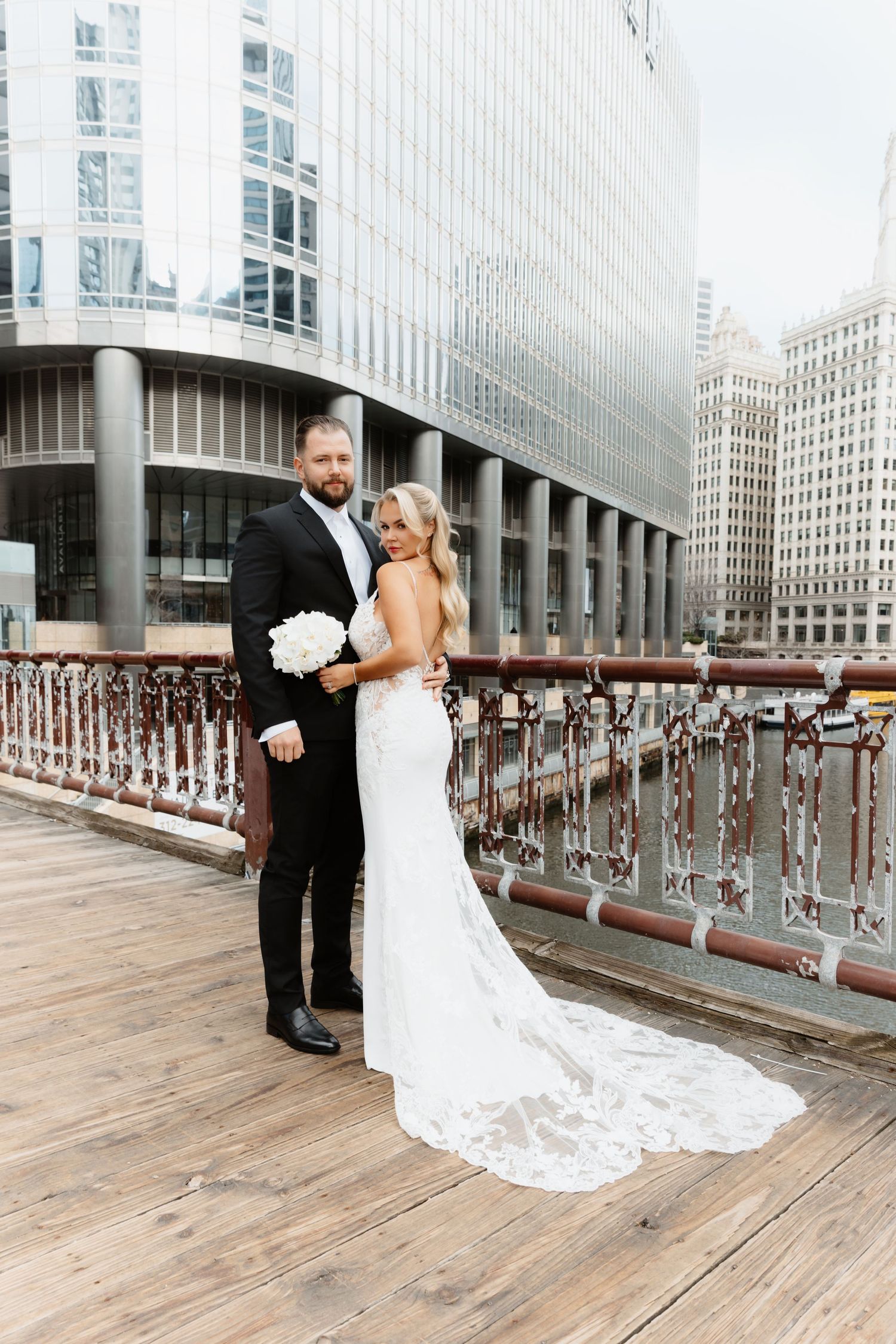 Bride in lace gown with long train and groom on Chicago Riverwalk bridge.
