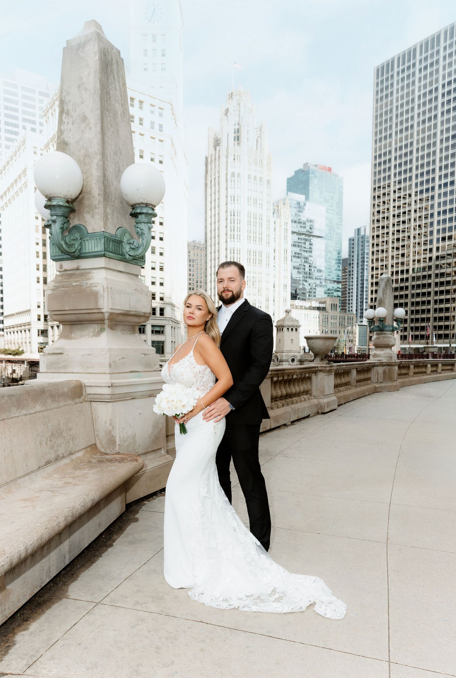 Bride and groom pose elegantly on Chicago bridge with Wrigley Building and skyline behind them.