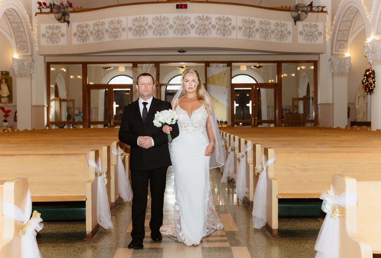 A bride in a white lace wedding gown walks the church aisle escorted by a man in a black suit.
