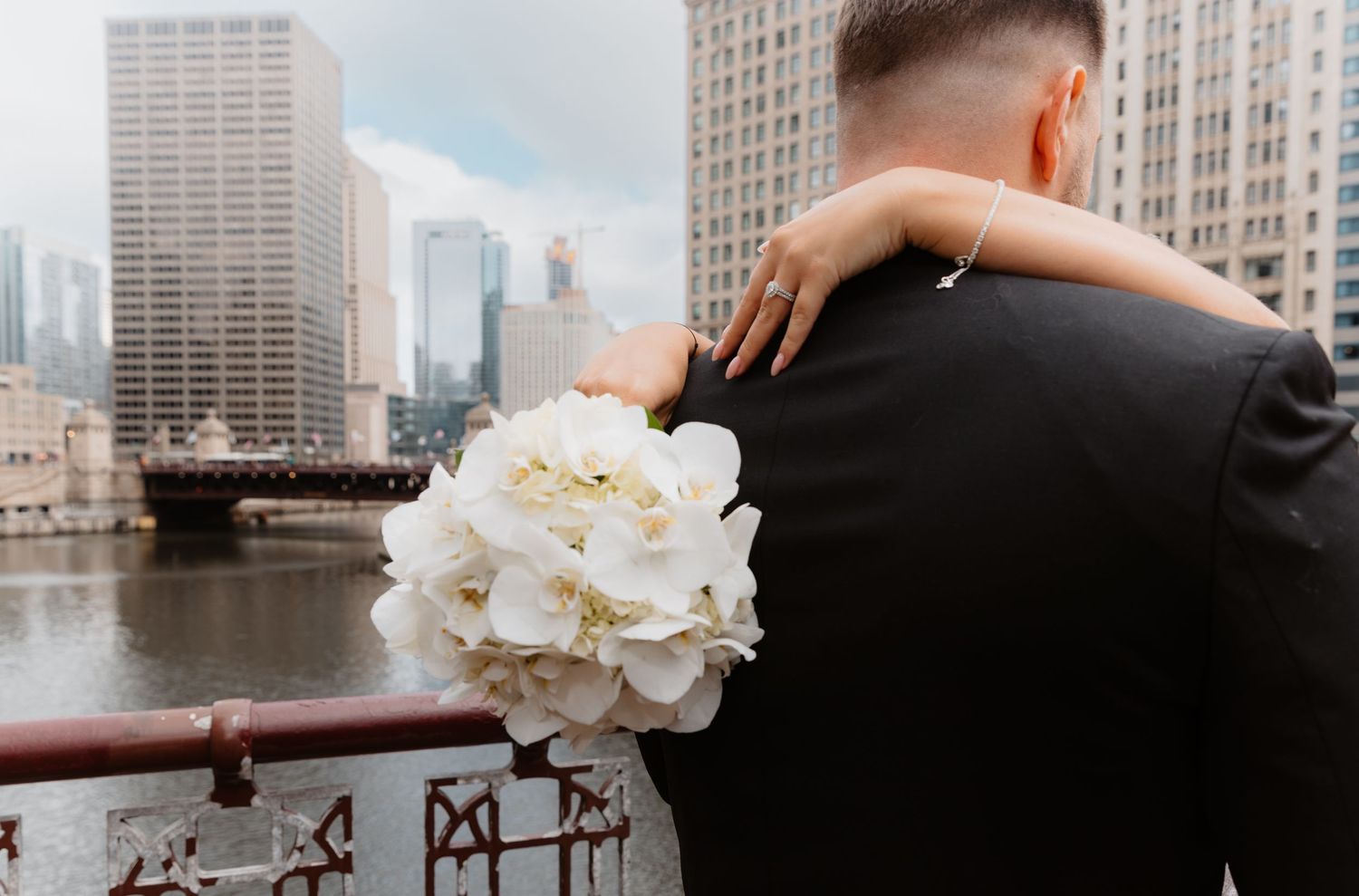 Bride holding white orchid bouquet embraces groom on Chicago River bridge with city skyline backdrop.