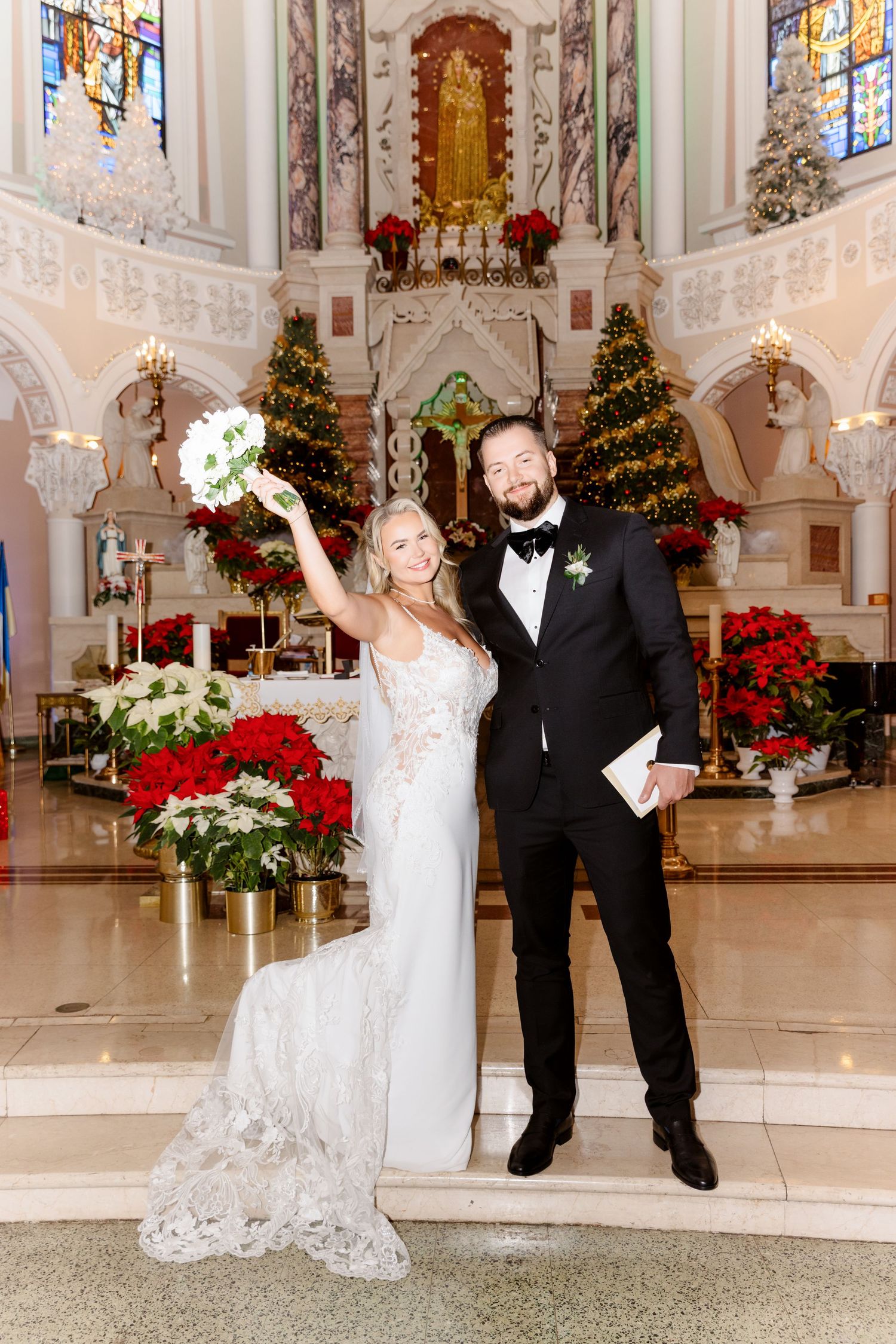 Bride and groom pose joyfully at church altar adorned with Christmas trees and red poinsettias after winter wedding ceremony.