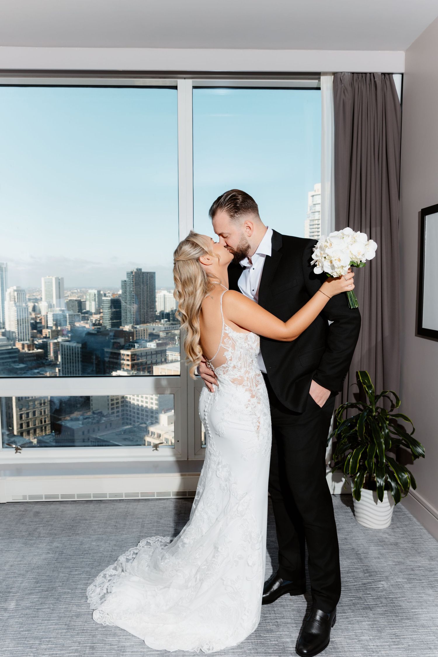 Bride and groom kiss romantically in a bright hotel room overlooking a beautiful city skyline.