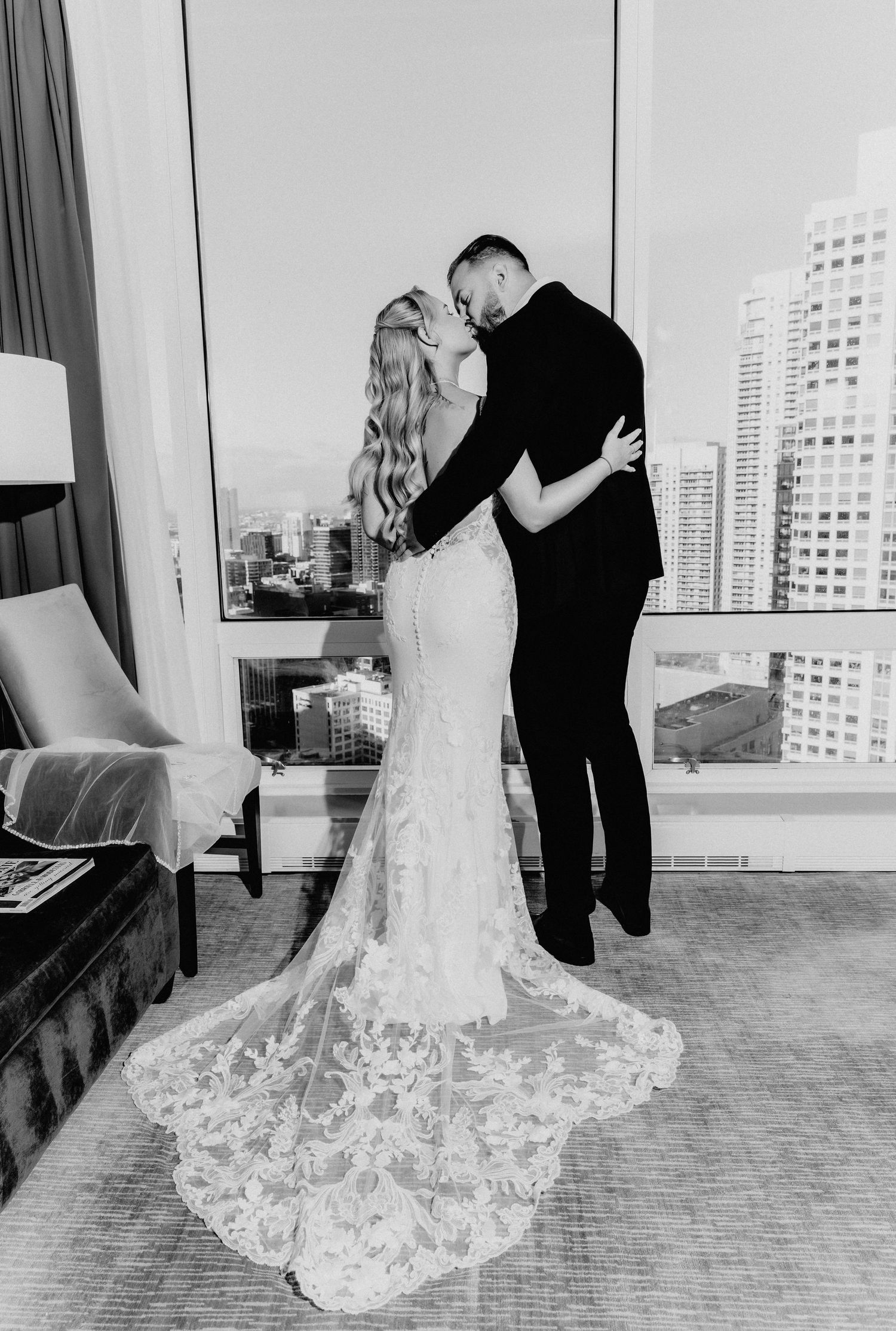 Black and white photo of newlyweds embracing in lace mermaid wedding gown with city skyline backdrop.