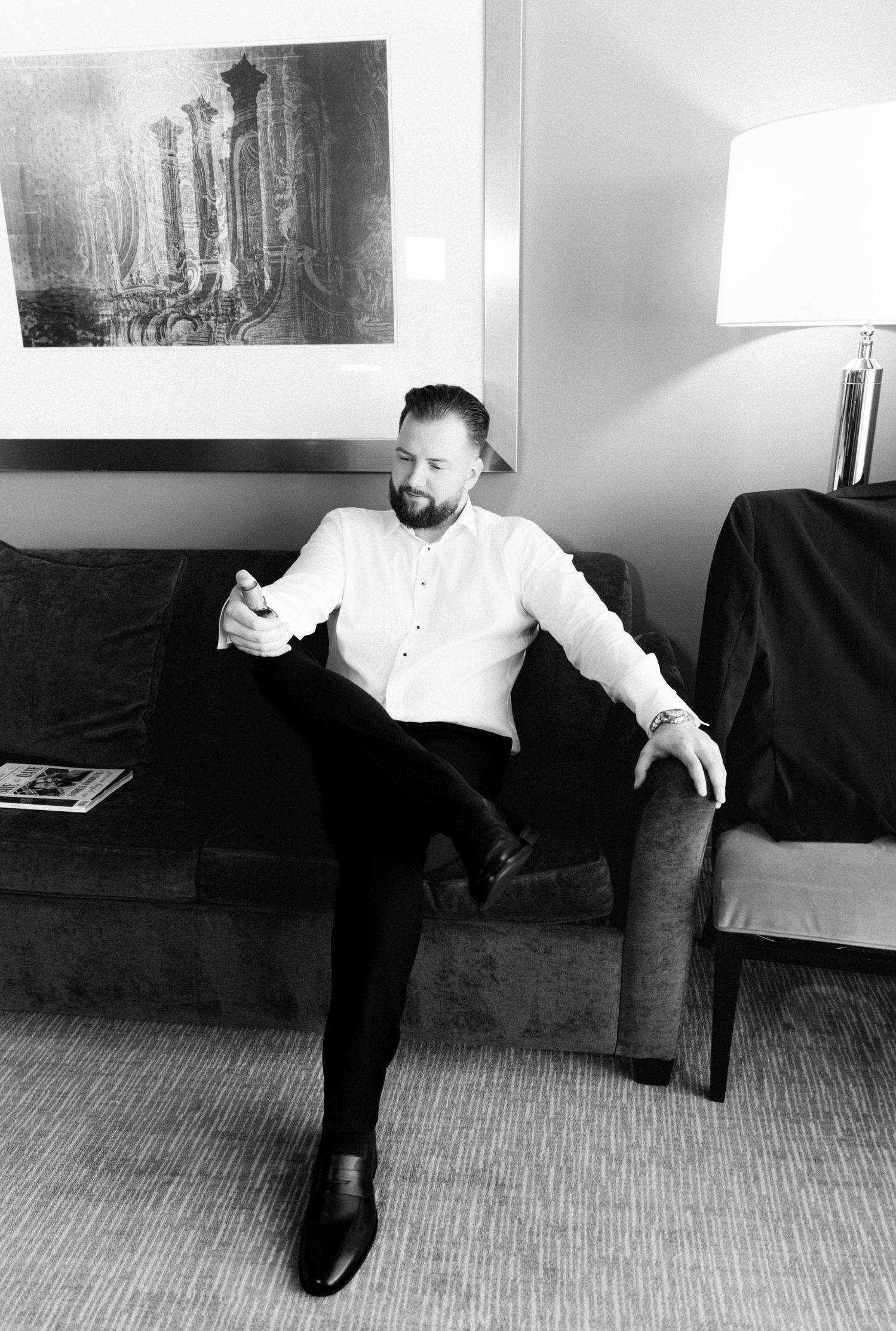 Groom in white dress shirt sits relaxed on sofa in black and white pre-wedding portrait.