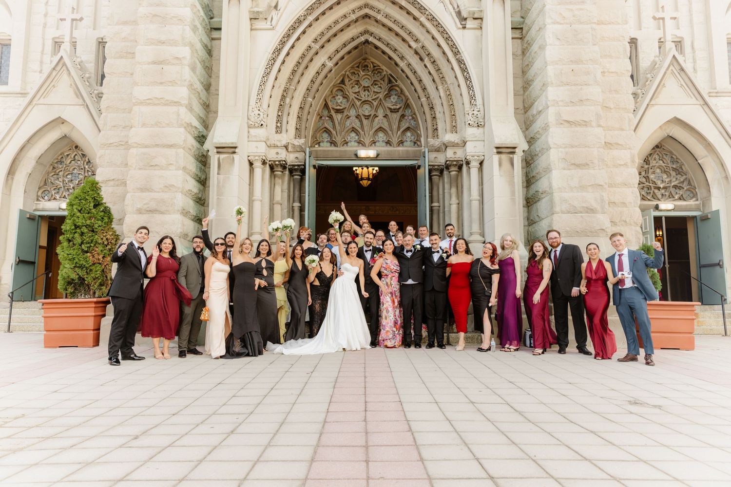 Wedding party celebrates with raised bouquet outside grand Gothic stone cathedral.