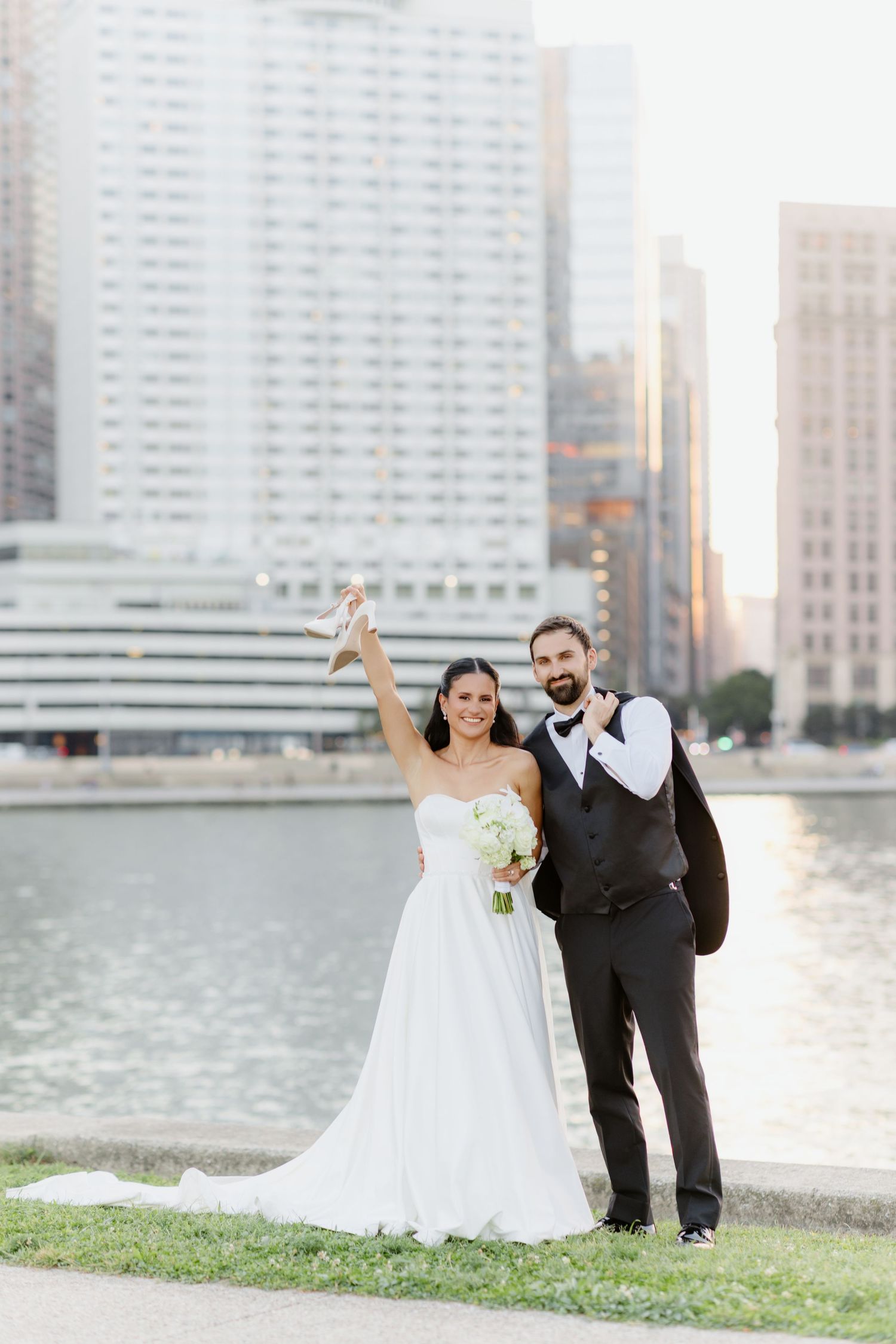 Bride and groom smile together on urban street at dusk, she in white gown holding white floral bouquet.
