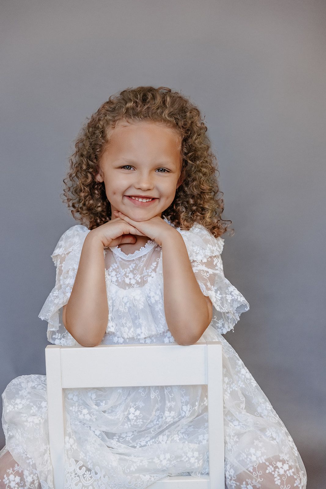 Smiling young girl with curly hair wearing a white lace dress, posing with chin on hands on a white chair.