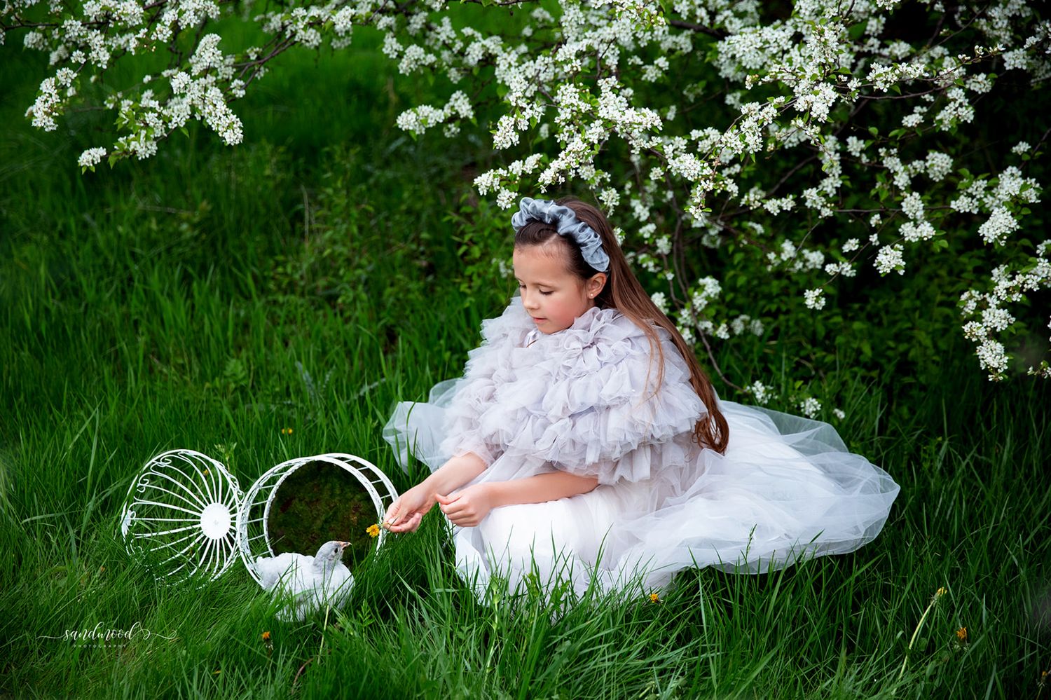 Young girl in white feathered dress sits in green grass near blooming white flowers, holding a wire basket.