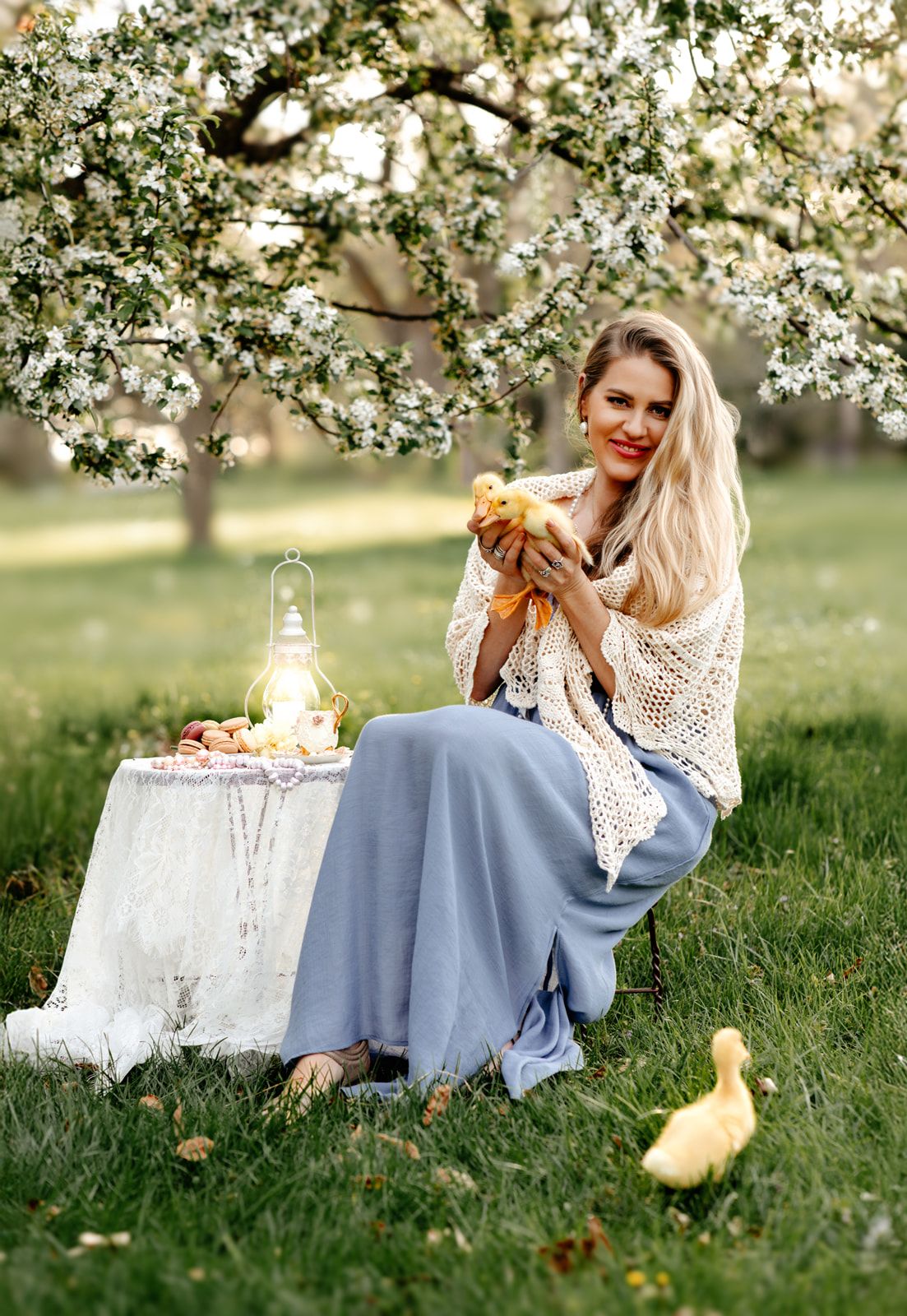Smiling blonde woman in blue skirt holding ducklings under blooming tree in spring garden with lantern.