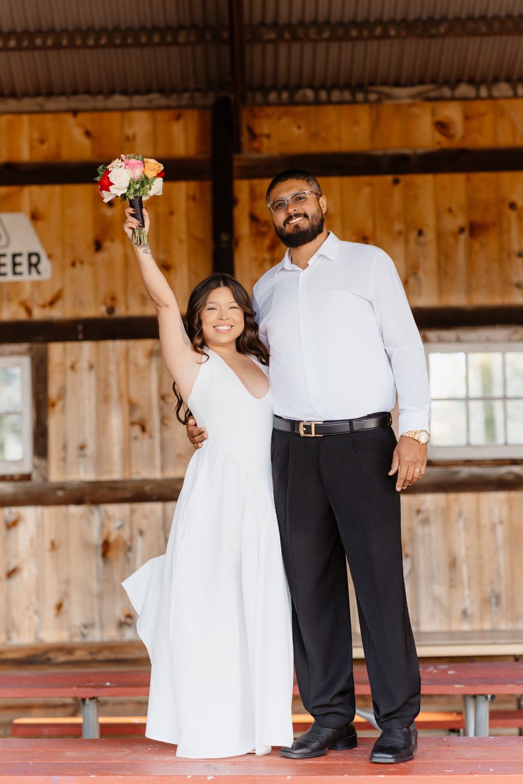 Joyful newlyweds pose in a rustic barn, bride in white dress raising bouquet, groom in white shirt and black pants.